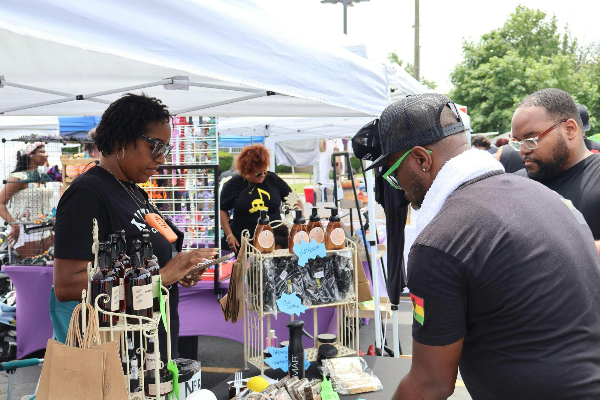 A vendor tent features body care products. A vendor uses their device to ring up customers while a group of attendees look at the items for sale.