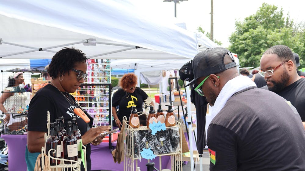 A vendor tent features body care products. A vendor uses their device to ring up customers while a group of attendees look at the items for sale.