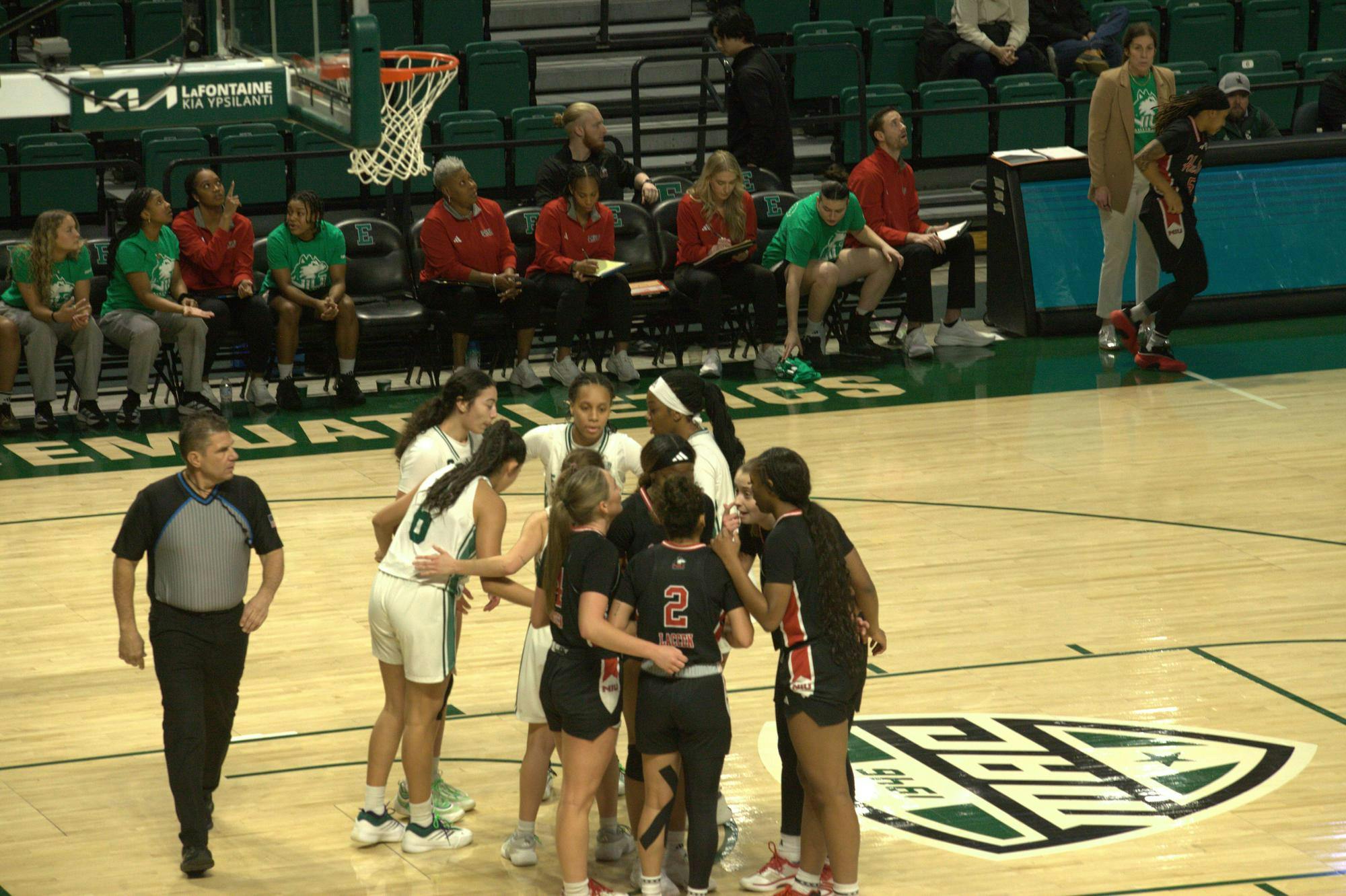 Easter Michigan and Northern Illinois Women's Basketball teams huddle in separate circles on court.