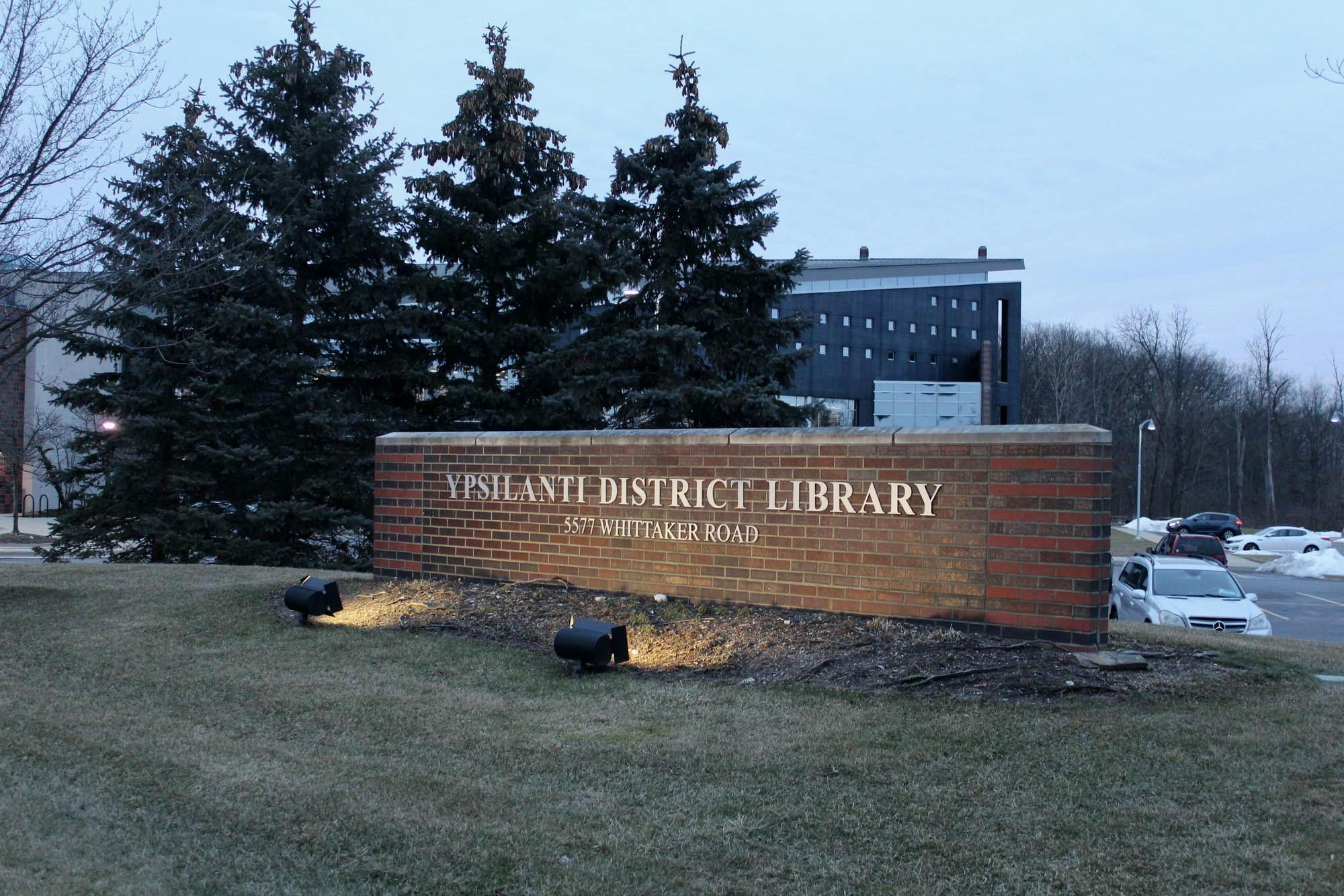 A sign made of brown and red bricks in front of several pine trees. The sign has two lines of text. The first line says, "Ypsilanti District Library," and the second says, "5577 Whittaker Road." The sign is outside, and the sky overhead is dusky.