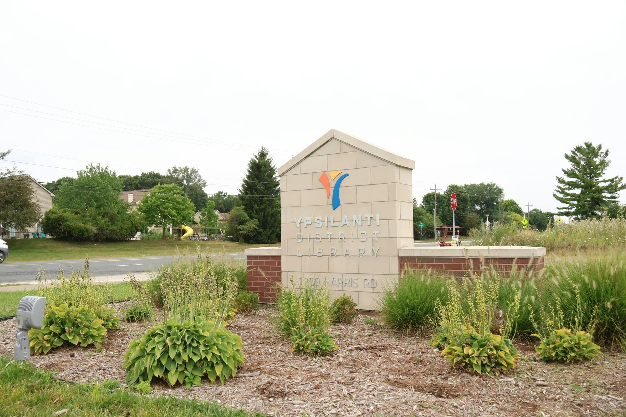 A sign made of pale bricks with the Ypsilanti District Library logo and 1900 Harris Road on it . It is surrounded by small bushes.