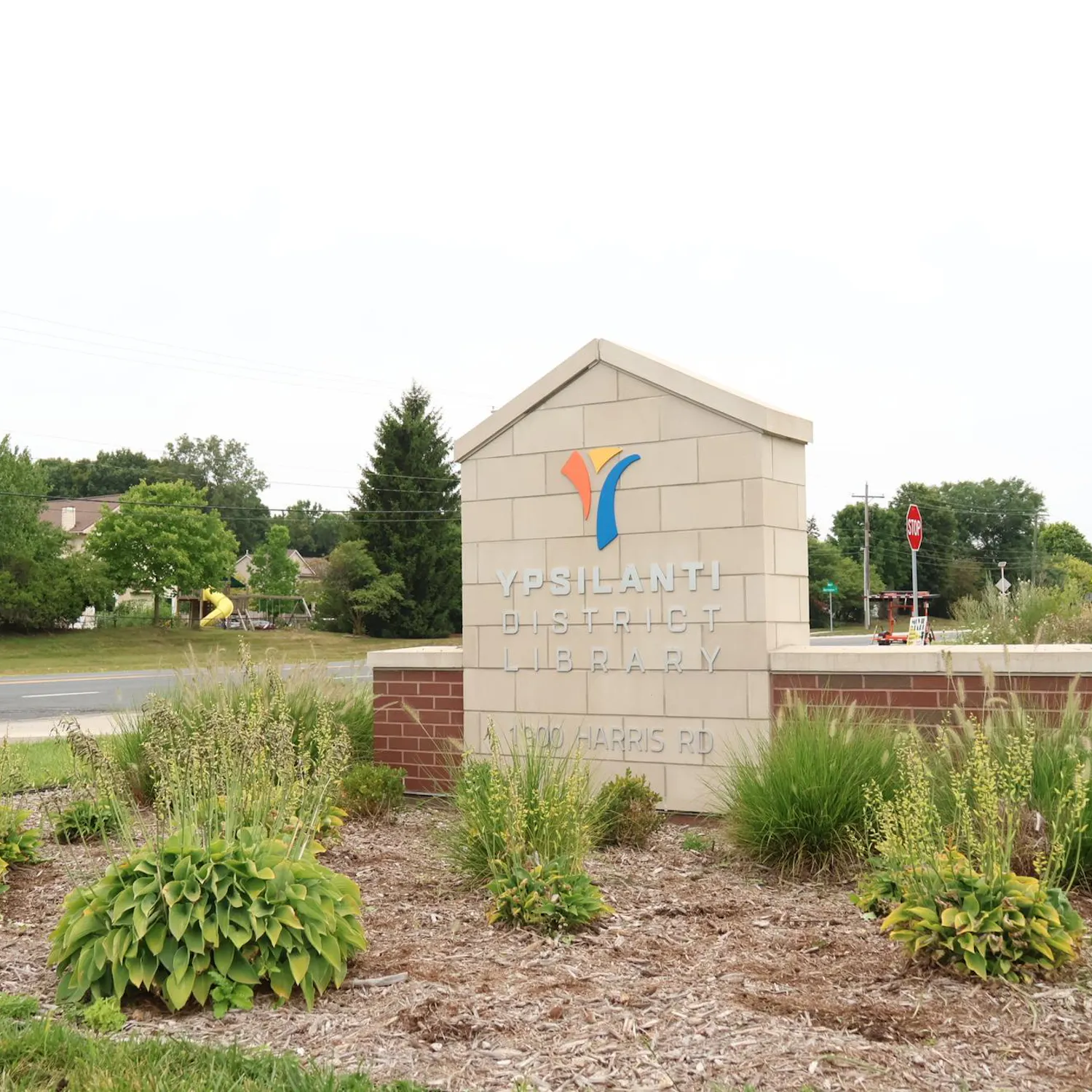 A sign made of pale bricks with the Ypsilanti District Library logo and 1900 Harris Road on it . It is surrounded by small bushes.