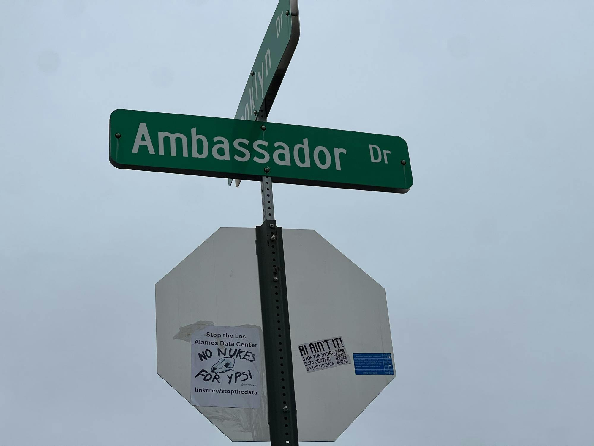 The back of a stop sign. A flyer stuck to the back says "No nukes for Ypsi" with a drawing of an animal skull and a link to the Stop the Data Center advocacy group Linktree page. The sticker next to it says "AI ain't it!" and is also attributed to Stop the Data Center.