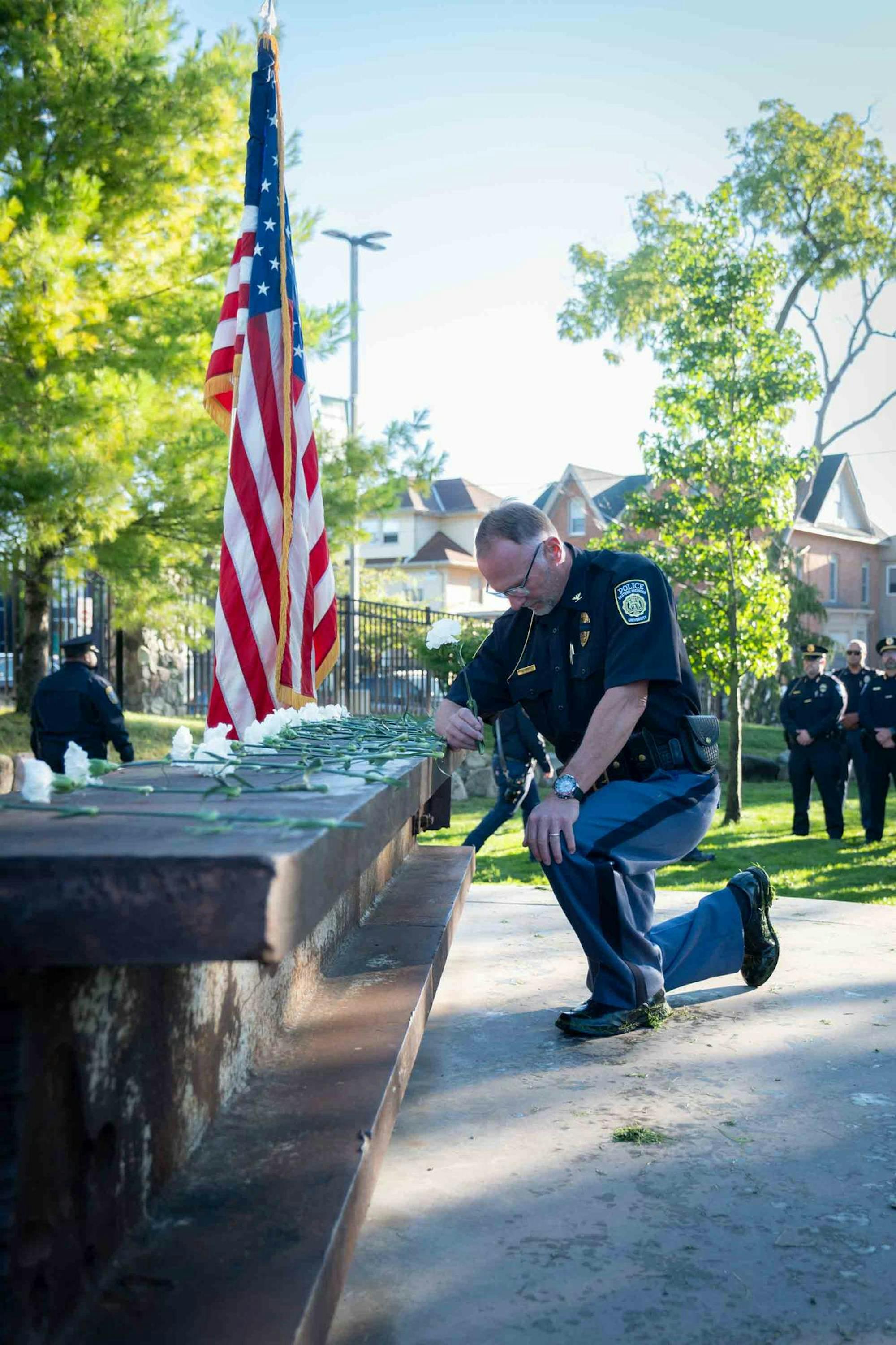 Matthew Lige, in a police uniform, kneels in front of a memorial comprised of a recovered steel beam from the South Tower of the World Trade Center. The beam is covered with white carnations. There is an American flag and more officers in the background.
