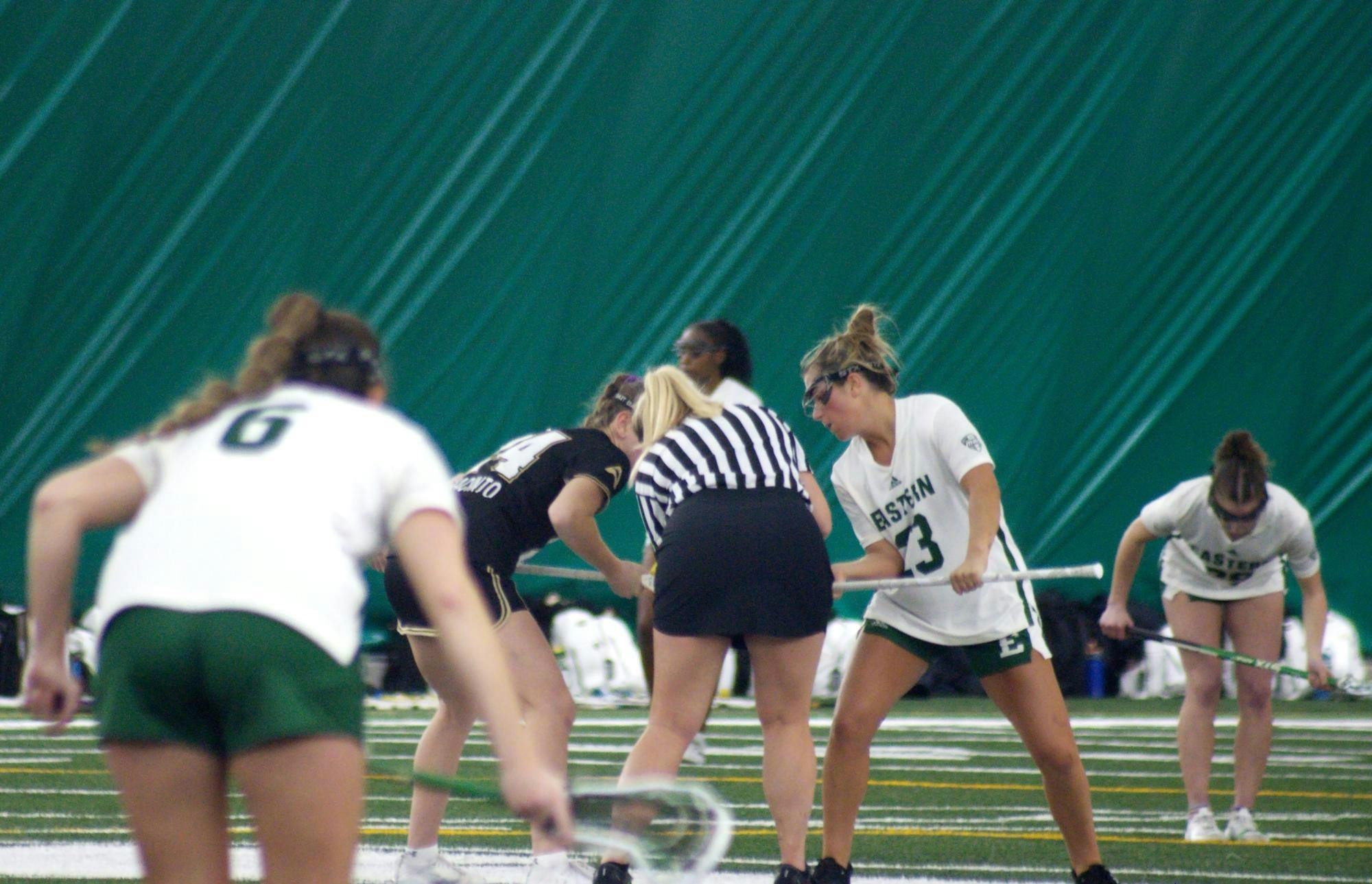 Two Lacrosse players in white Eastern Michigan jerseys stand off to the sides while an Eastern Michigan player and Lindenwood player in a black jersey stand at the center with a referee between them.