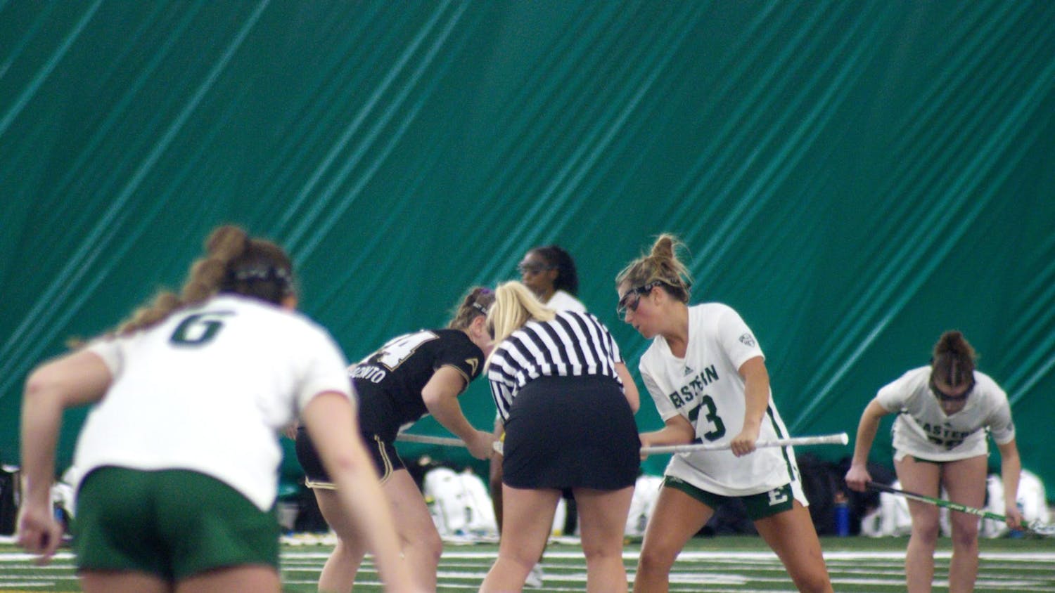 Two Lacrosse players in white Eastern Michigan jerseys stand off to the sides while an Eastern Michigan player and Lindenwood player in a black jersey stand at the center with a referee between them.
