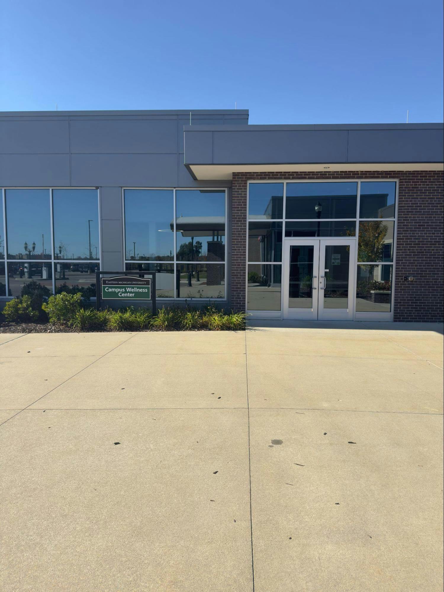 The front entrance of a brick building with big windows and glass double doors. A dark green sign in front of the building reads, "Campus Wellness Center" in white letters. 