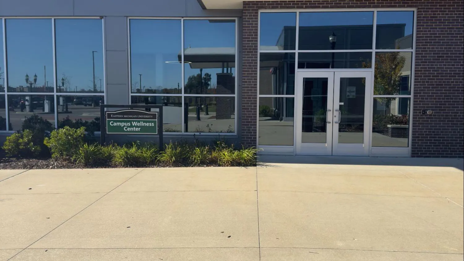The front entrance of a brick building with big windows and glass double doors. A dark green sign in front of the building reads, "Campus Wellness Center" in white letters.