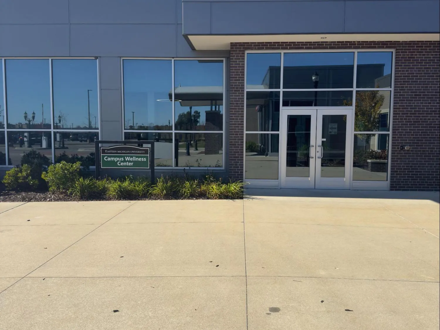 The front entrance of a brick building with big windows and glass double doors. A dark green sign in front of the building reads, "Campus Wellness Center" in white letters.