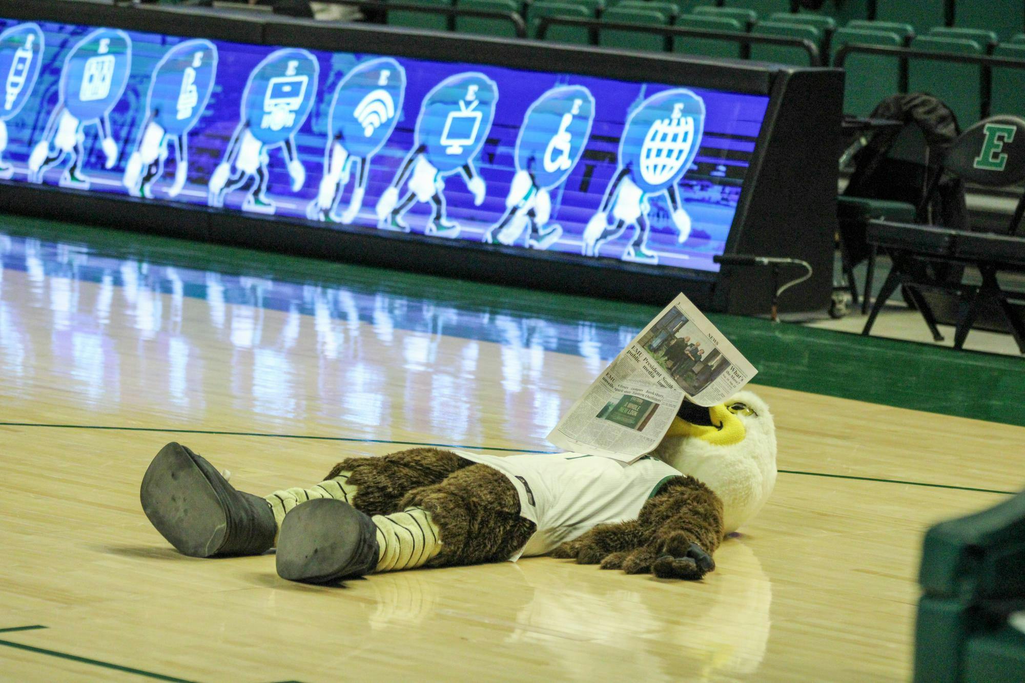 Swoop, EMU's mascot, is reading The Eastern Echo newspaper while lying on his back on the basketball court.