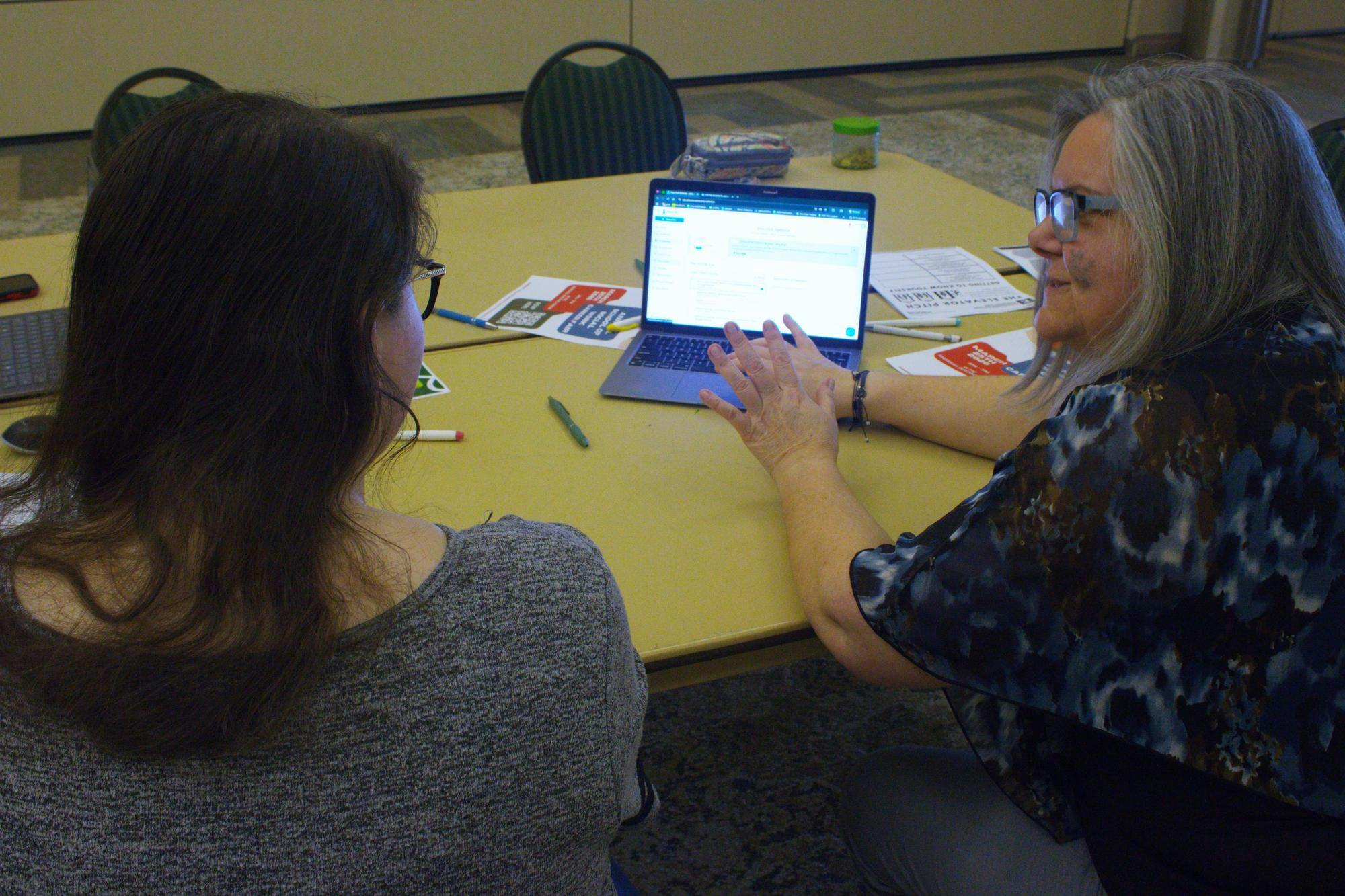 A student and an EMU faculty member sit facing a laptop that rests on a cluttered table.
