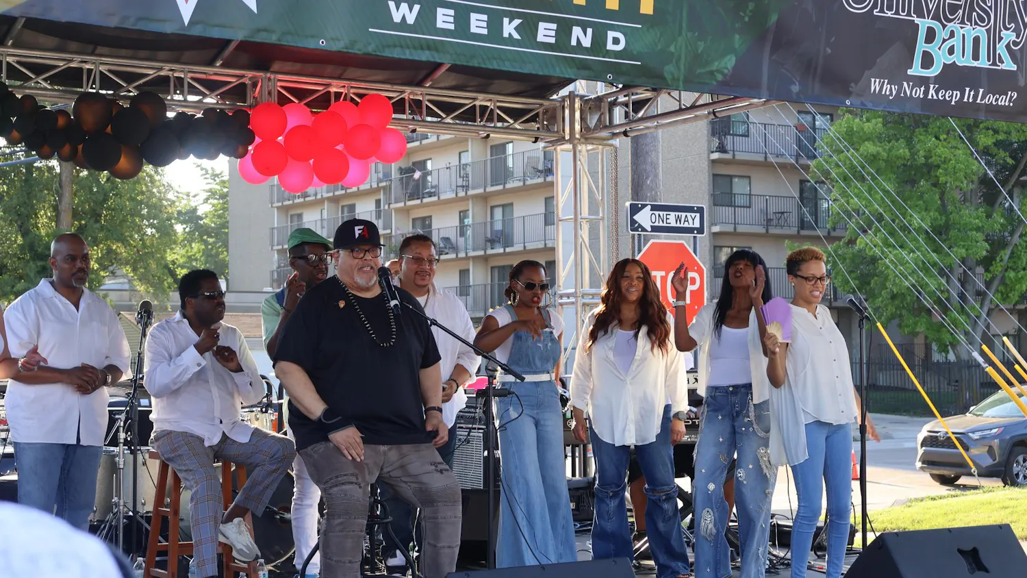 Fred Hammond sits on stage in front of a microphone in a snapback hat, black t-shirt and grey jeans with multiple background performers surrounding him in matching white tops and blue jeans.
