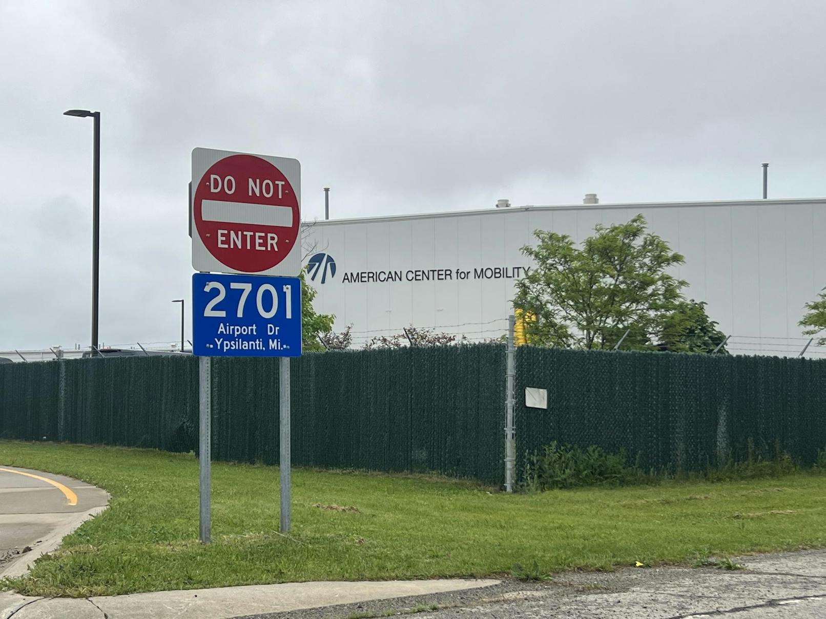 An address sign labled "2701 Airport Dr Ypsilanti Mi.," a wire fence, and a few trees frame the sign on the American Center for Mobility building. 