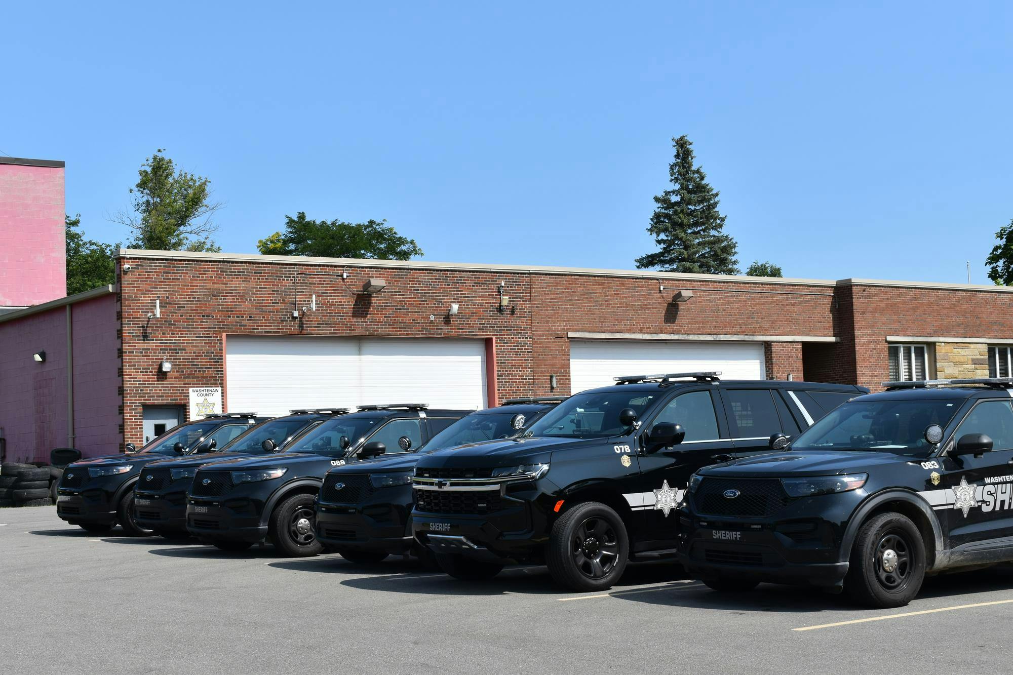 6 black police SUVs parked in front of a red brick building on a sunny day.