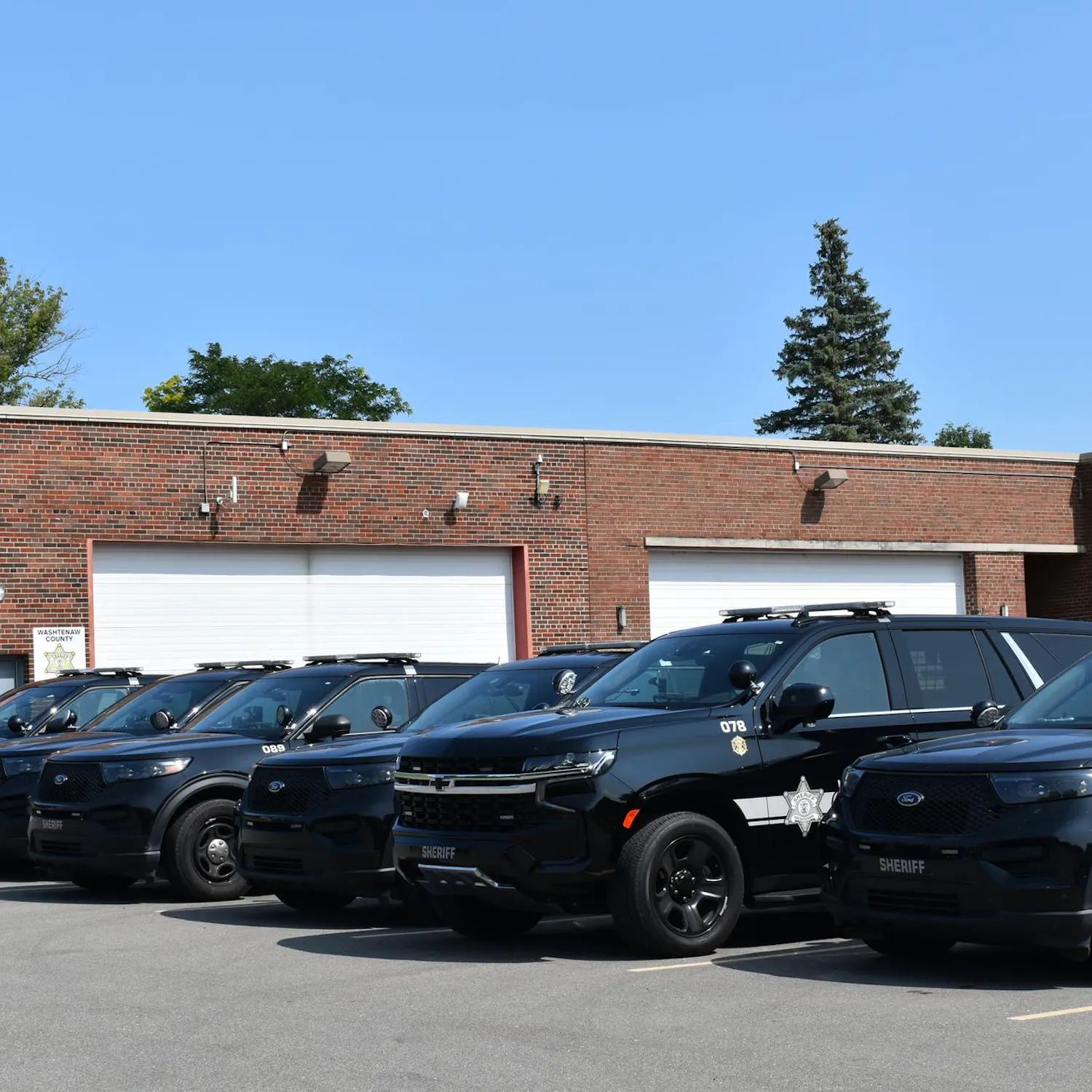 6 black police SUVs parked in front of a red brick building on a sunny day.