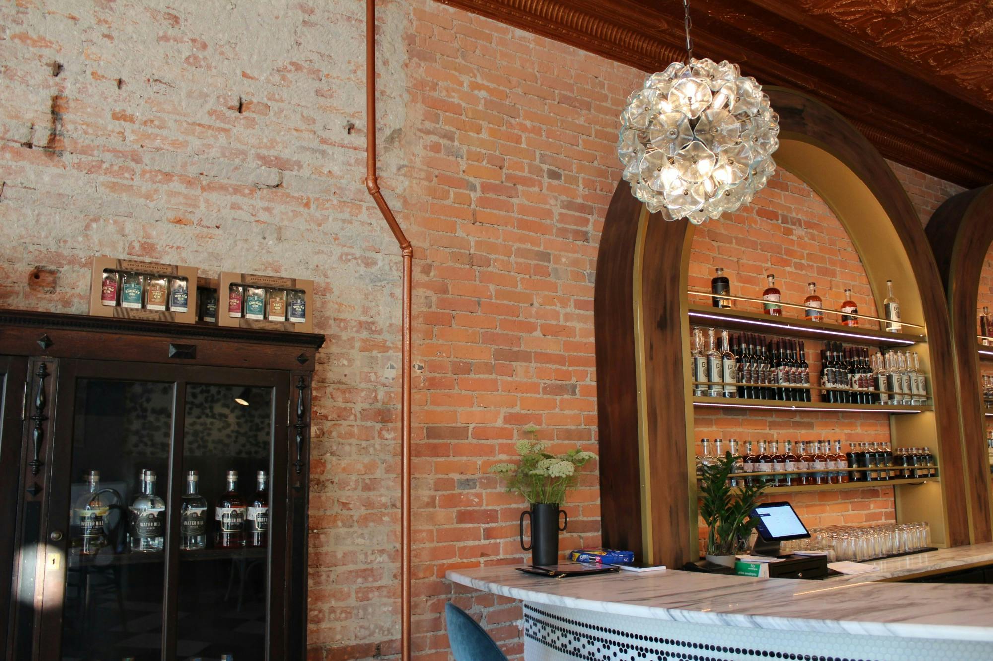 The bar area of the Ypsi Cocktail Club includes a counter, several shelves stocked with beverages, and a beautiful crystal lamp hanging overhead. All these details are brought together by the red brick wall of the establishment.