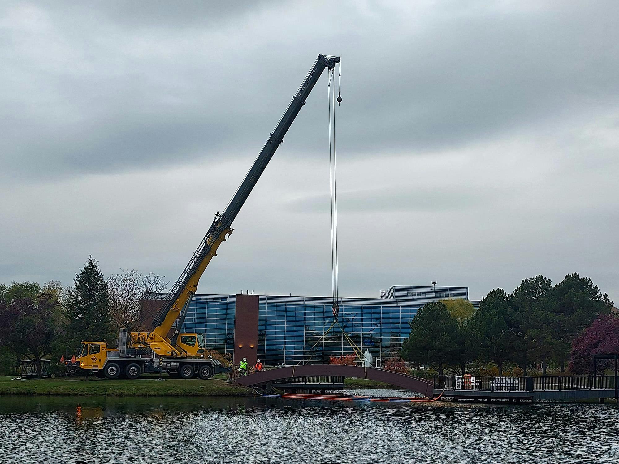 A large crane is tied to an arched wooden bridge, ready to lift it away from its position over the pond.