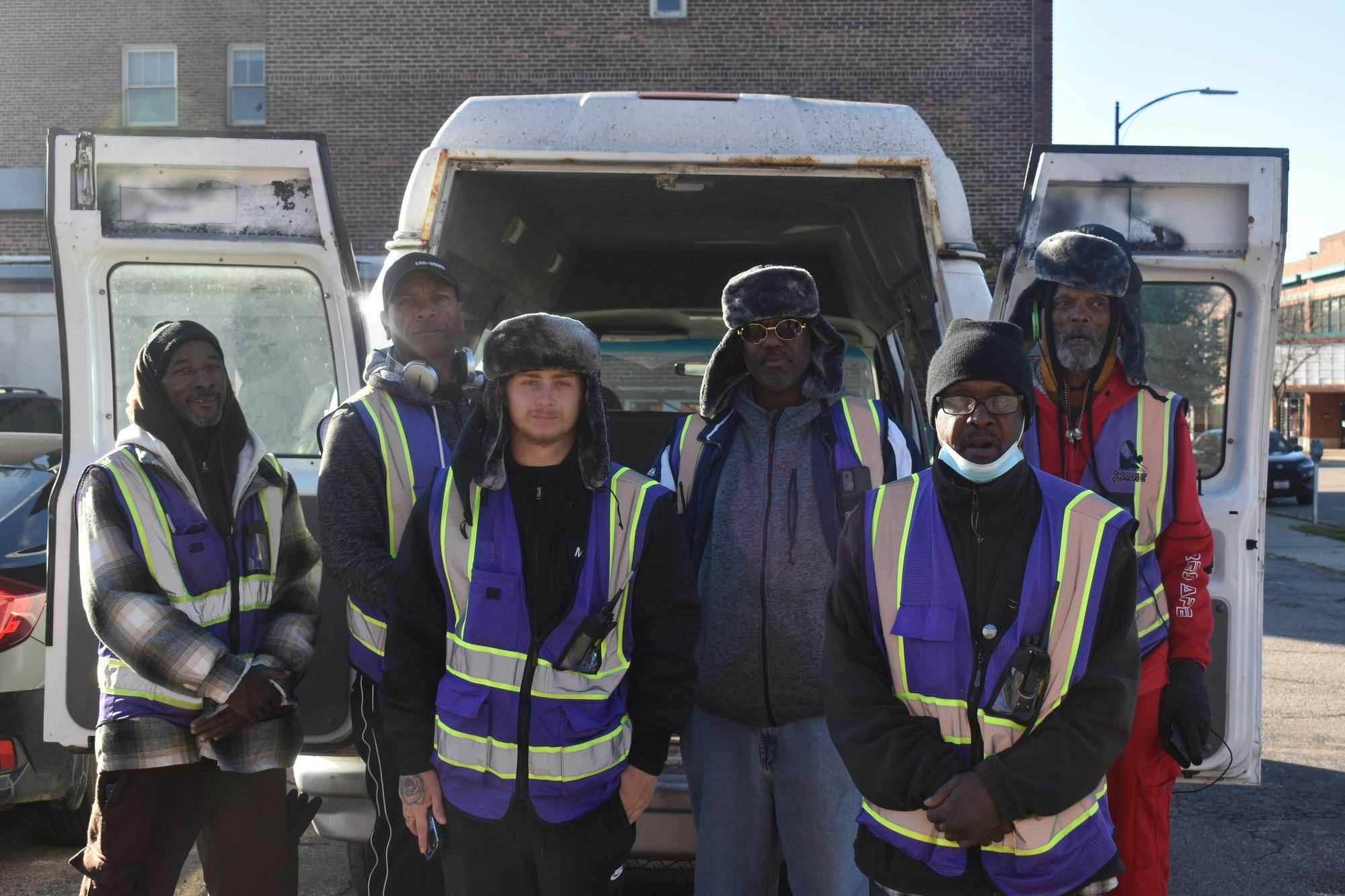 A crew of 6 men in purple vests from SHINE standing outside a building and behind a van they use.
