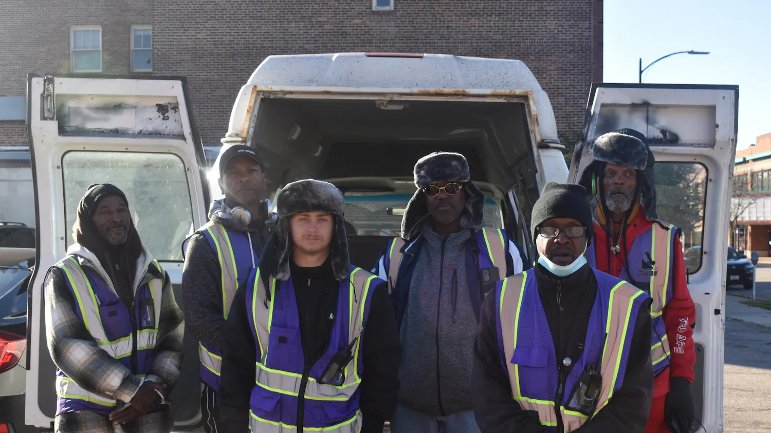 A crew of 6 men in purple vests from SHINE standing outside a building and behind a van they use.