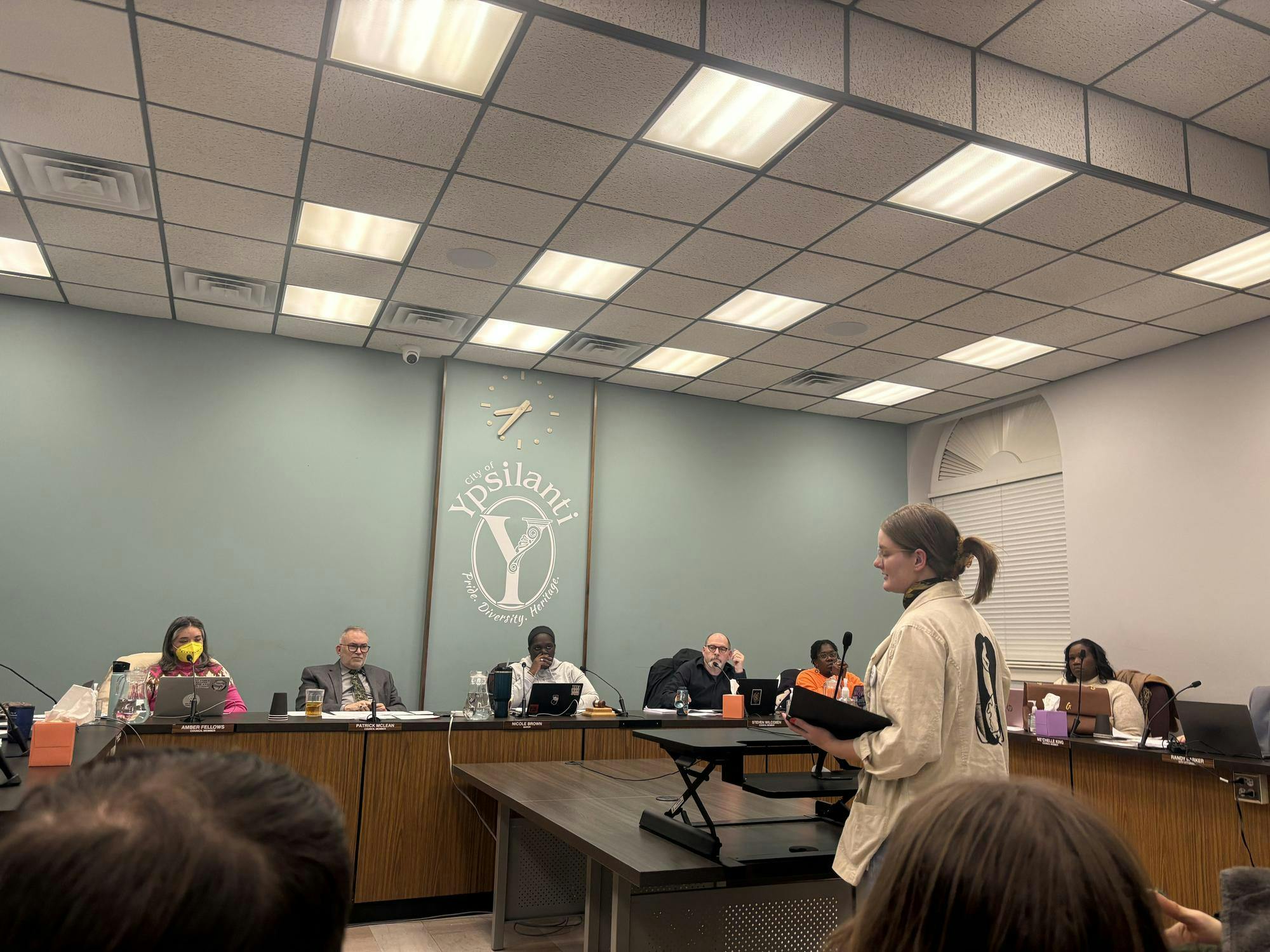 Mackenzie Stolzenburg, dressed in a cream-colored jacket, stands in front of the Ypsilanti City Council members, reading from a black notebook. Council members are seated in front of a blue wall that reads "City of Ypsilanti. Pride. Diversity. Heritage."