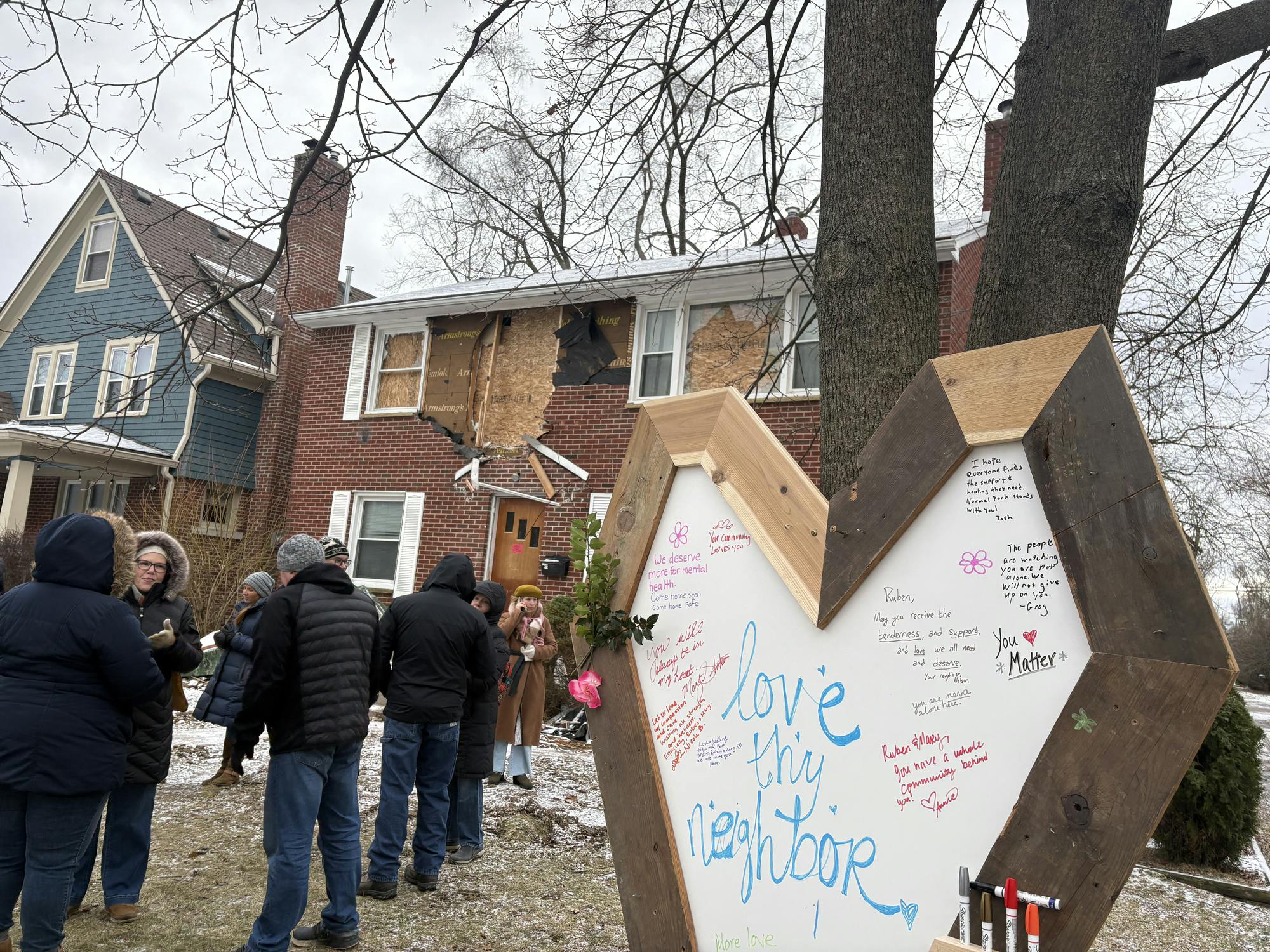 A large, hand-crafted heart-shaped sign with "Love thy neighbor" written in the center, along with other messages of support, stands in the yard of a damaged and condemned two-story brick house where several community members gather.