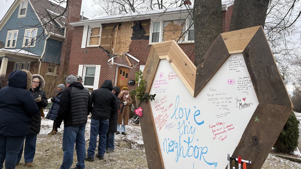 A large, hand-crafted heart-shaped sign with "Love thy neighbor" written in the center, along with other messages of support, stands in the yard of a damaged and condemned two-story brick house where several community members gather.