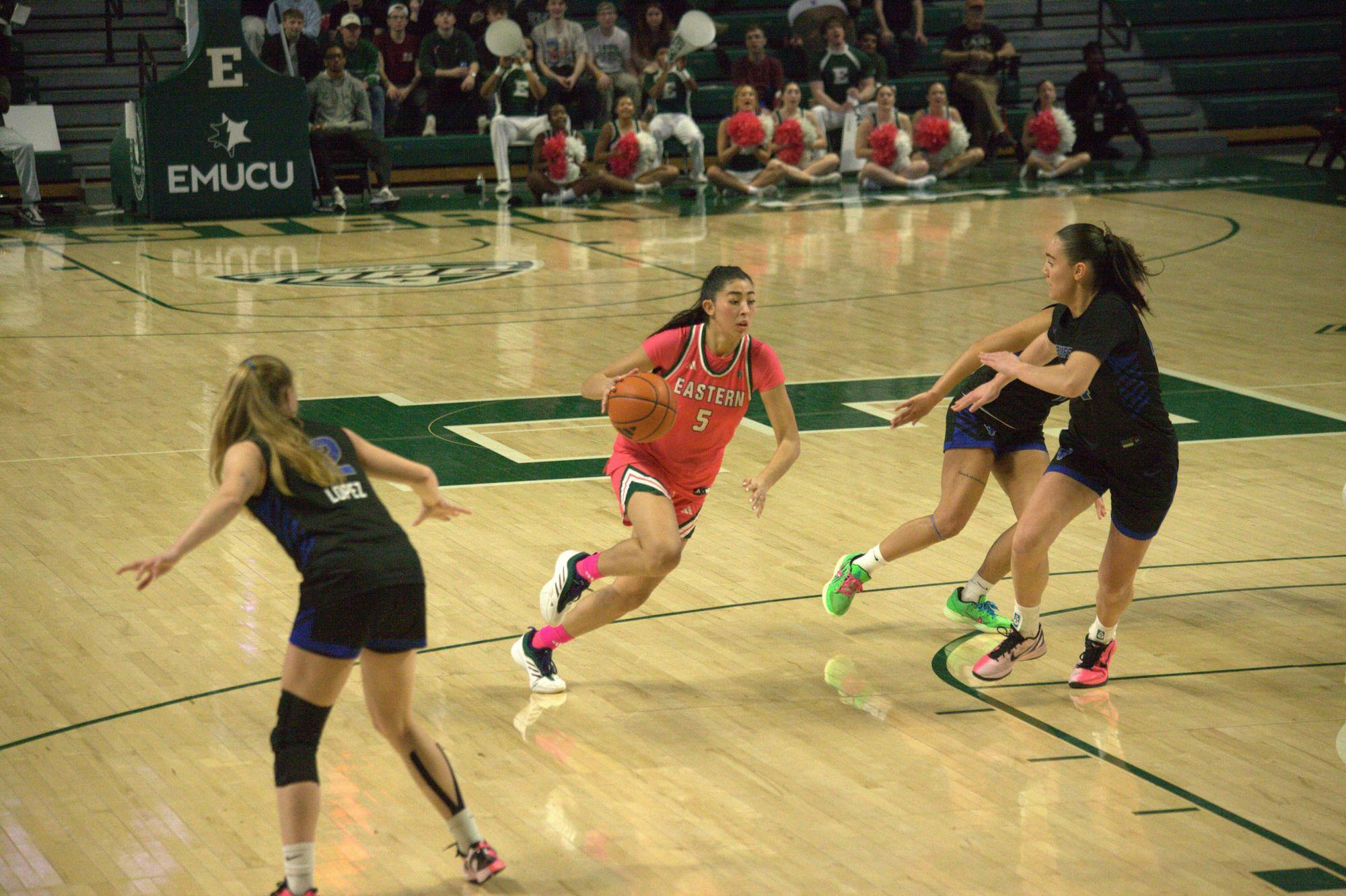 On an indoor court, a women's basketball player dribbles the ball in between members of the opposing team.