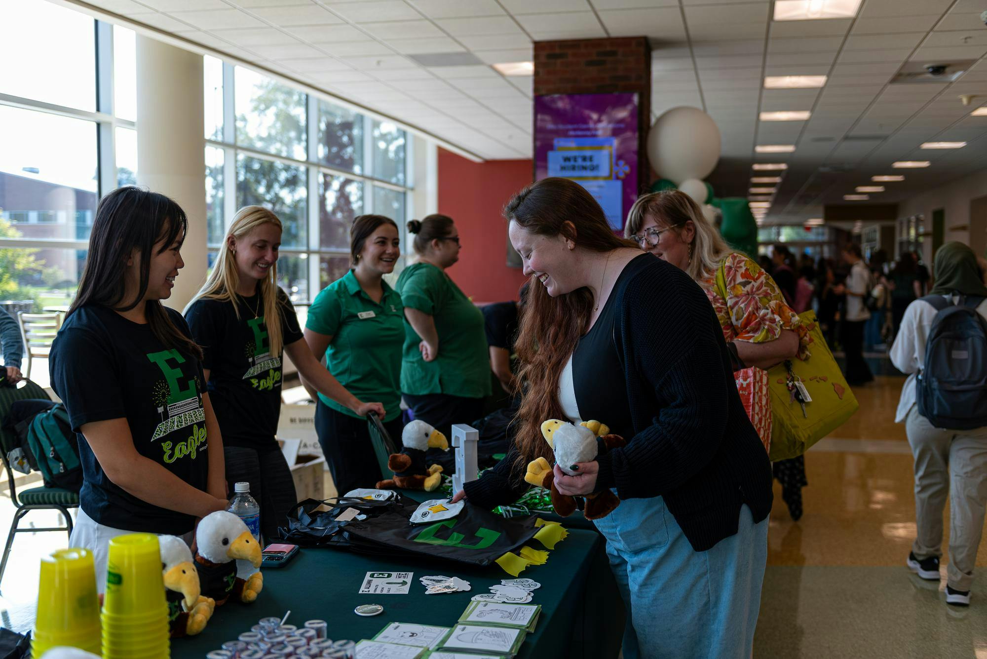 Hannah Fox holds a stuffed eagle and talks to people working a table in the Student Center. The table is covered with green swag items.