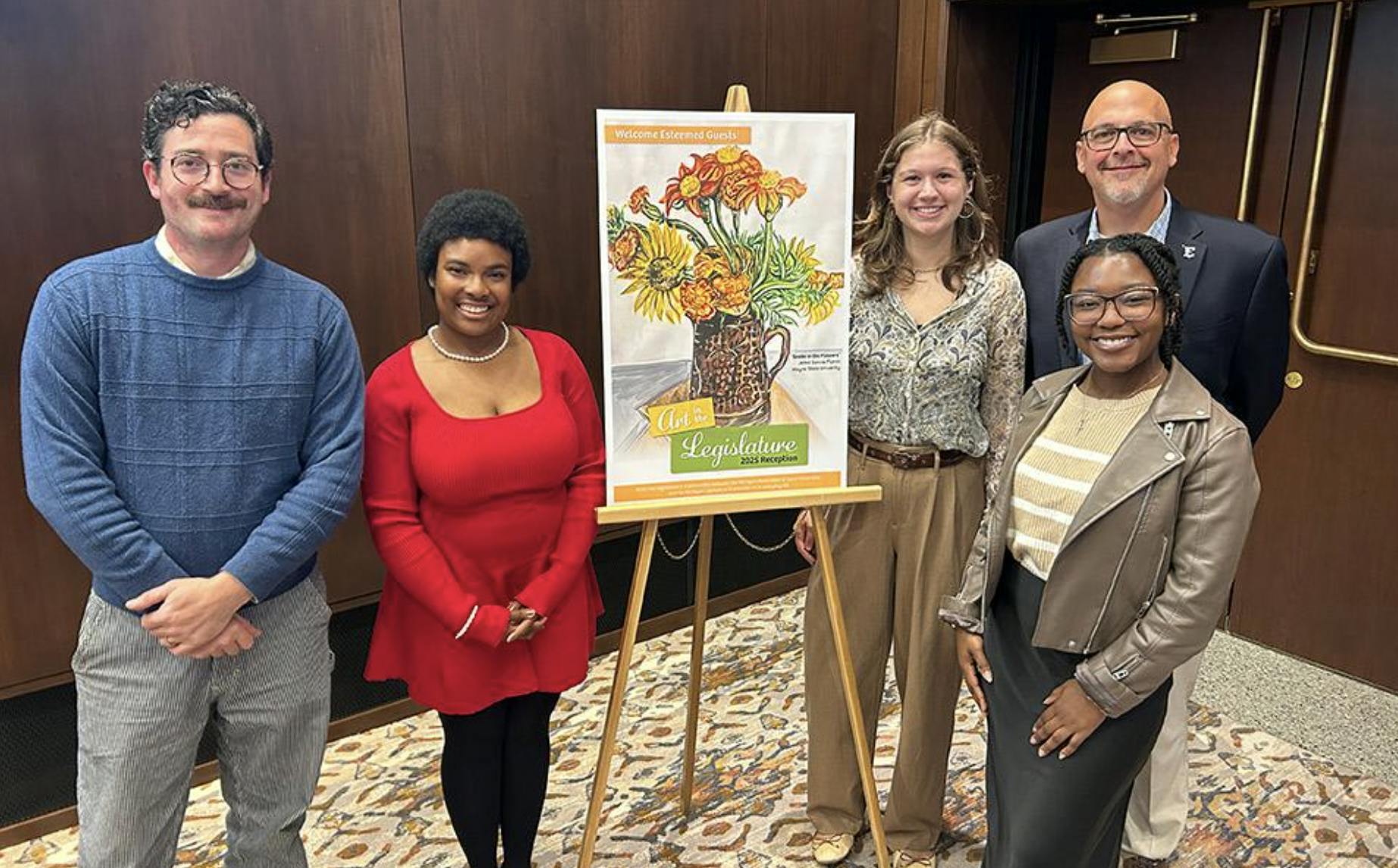 Five people smile and stand next to a sign that says "Art in the Legislature" displayed on an easel. 