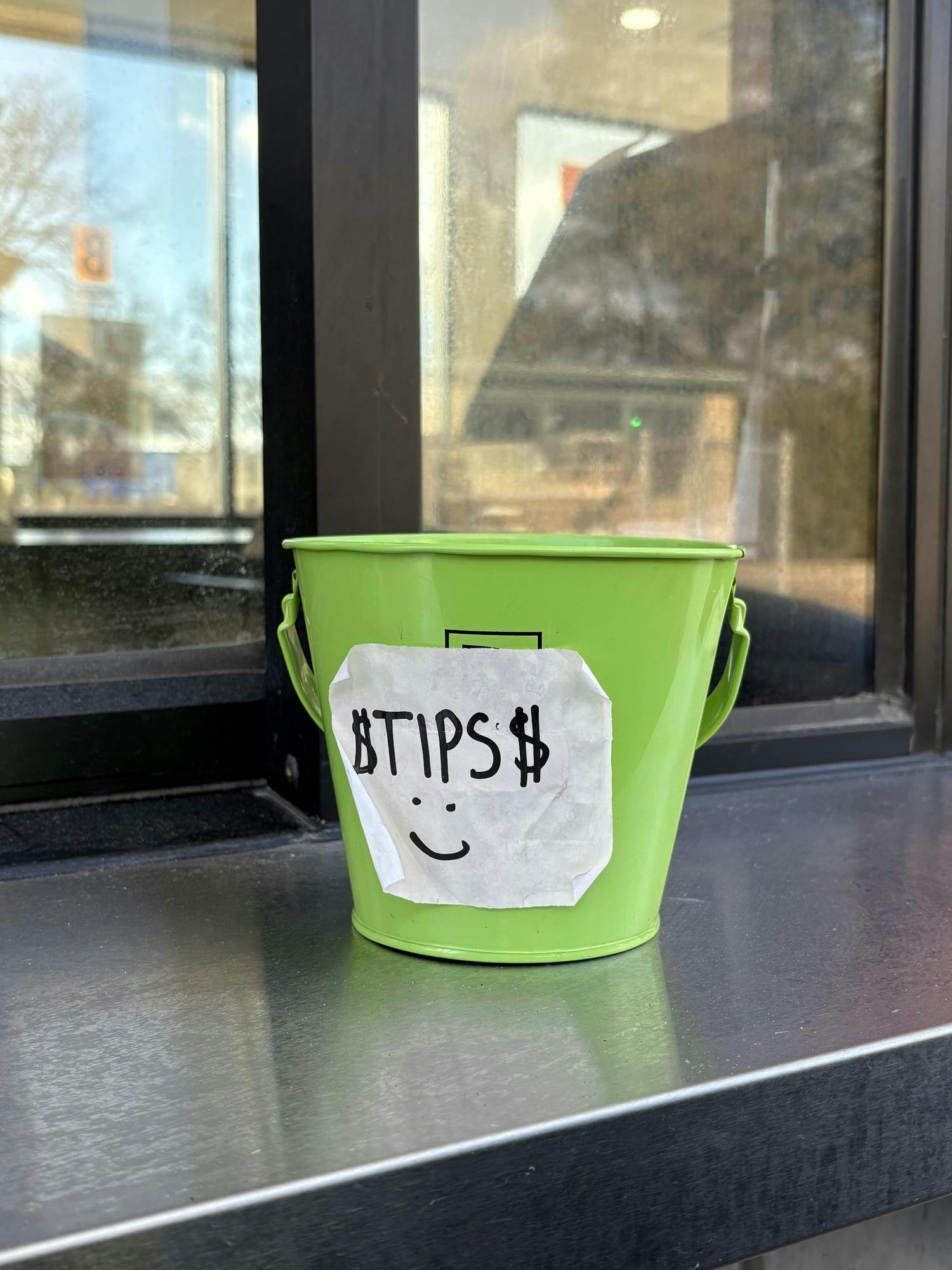 A bright green bucket sits on a metal ledge outside a drive-though window with a white sign taped on that says "Tips" with a smiley face. 