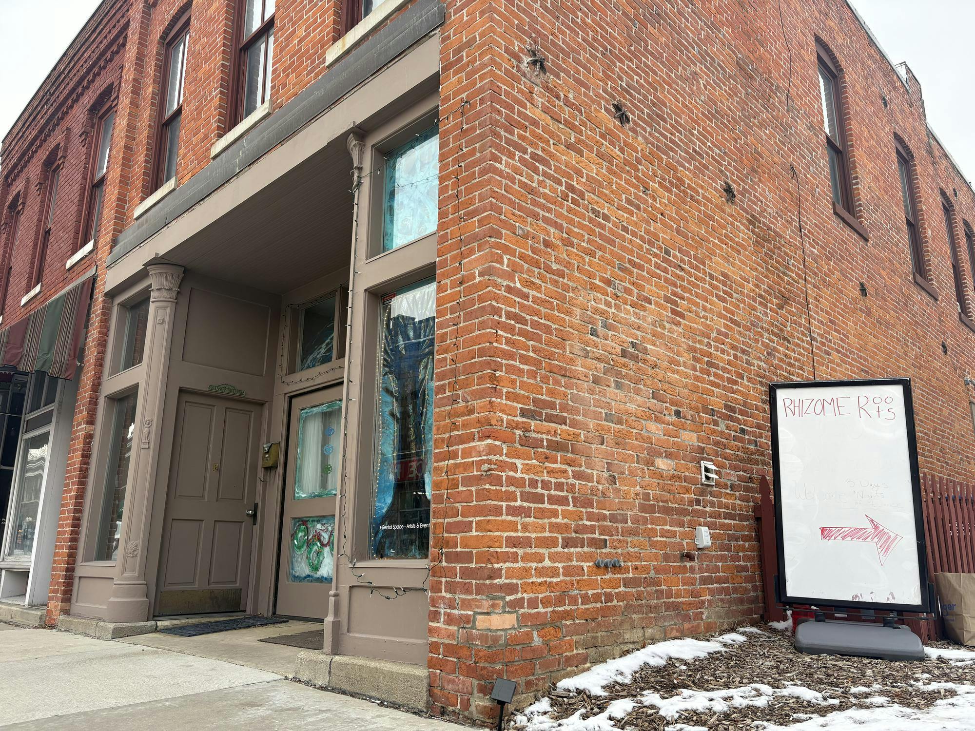 Corner of a historical brown brick building with brown trim and glass windows. To the right of the building is a sign that reads "Rhizome Roots" with an arrow pointing to the right.