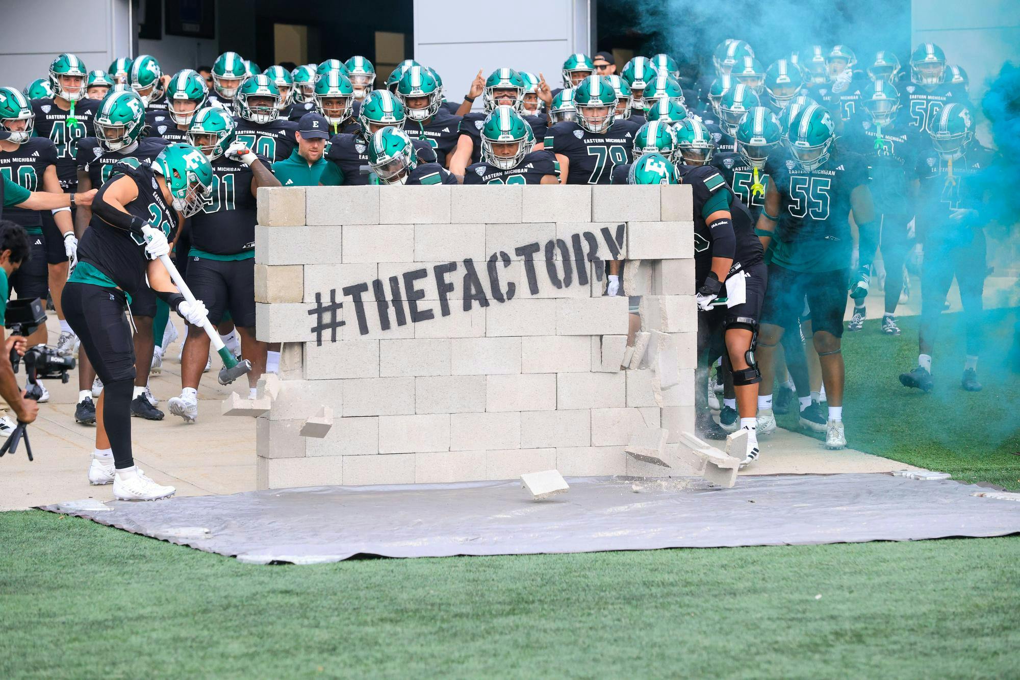 Using sledgehammers, teammates knock down a brick wall that says "The Factory."