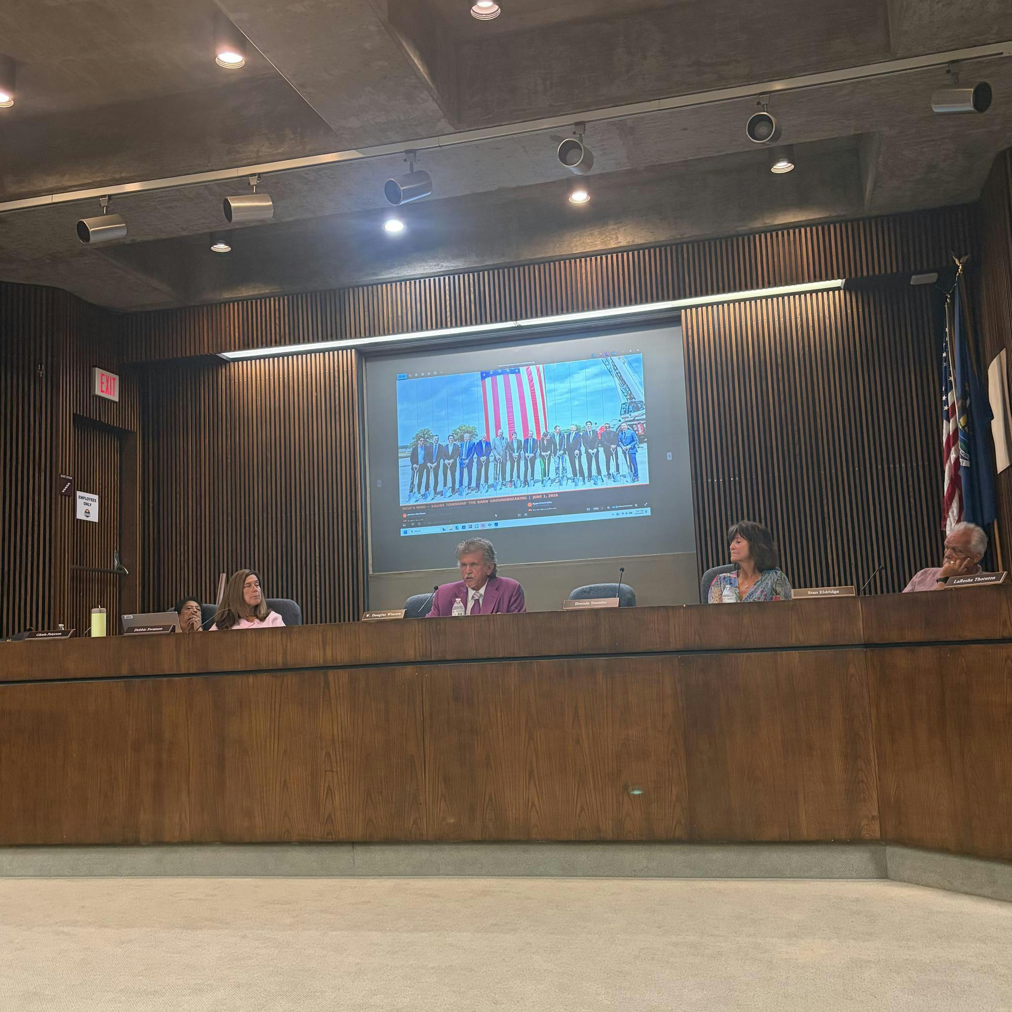 Members of the board sit at the wooden desk with a screen behind them.