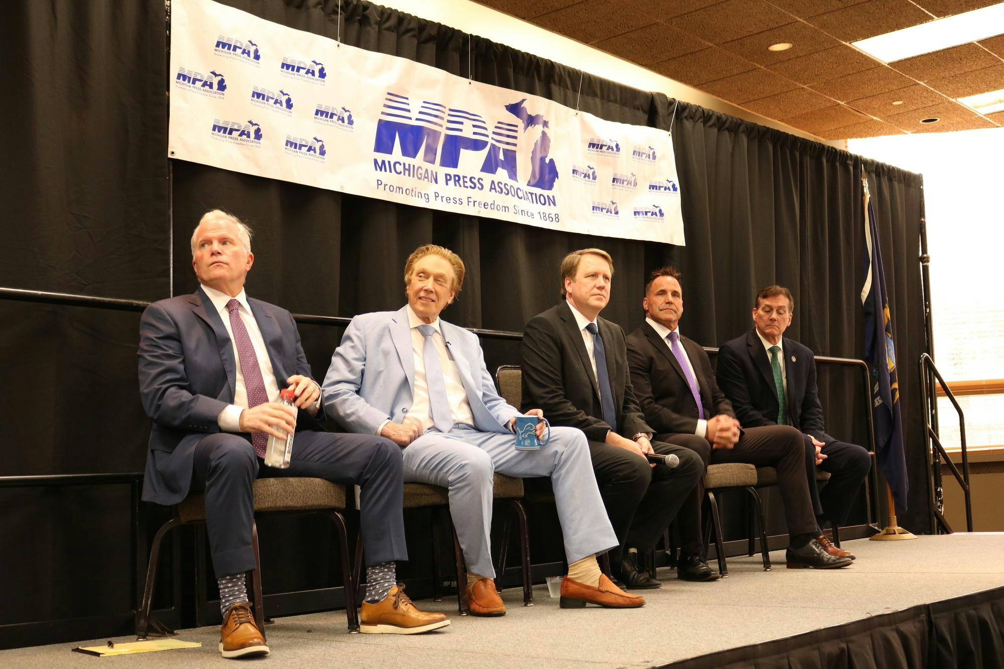 Five candidates sit on stage with a black curtain in the background and a banner that reads, "Michigan Press Association: Promoting Press Freedom Since 1868."