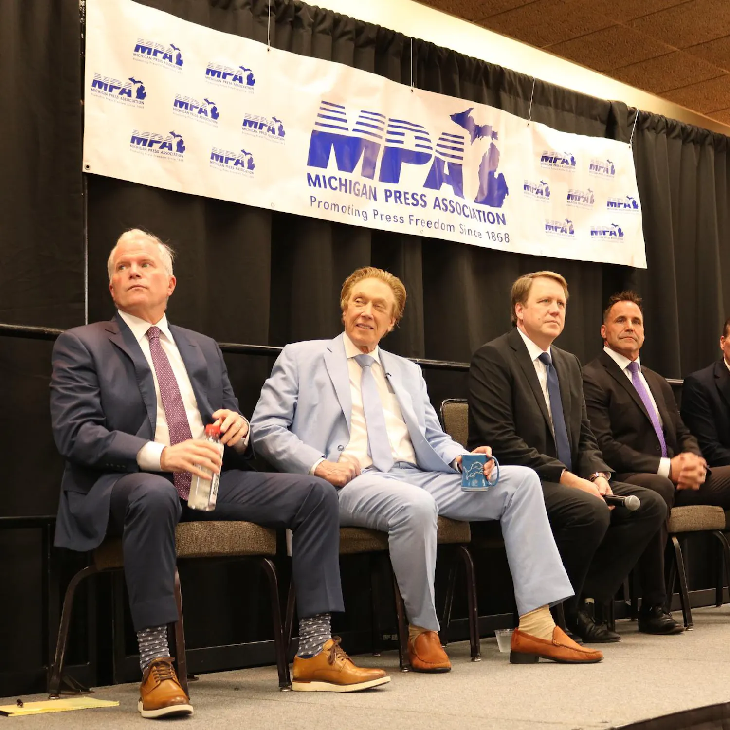 Five candidates sit on stage with a black curtain in the background and a banner that reads, "Michigan Press Association: Promoting Press Freedom Since 1868."