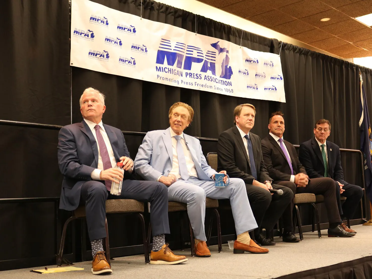 Five candidates sit on stage with a black curtain in the background and a banner that reads, "Michigan Press Association: Promoting Press Freedom Since 1868."