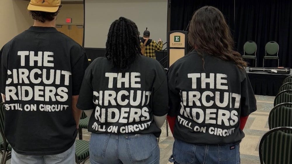 Three people stand in front of a stage with their backs to the camera. They wear the same black t-shirt with the words "The Circuit Riders, Still on Circuit" on the back.
