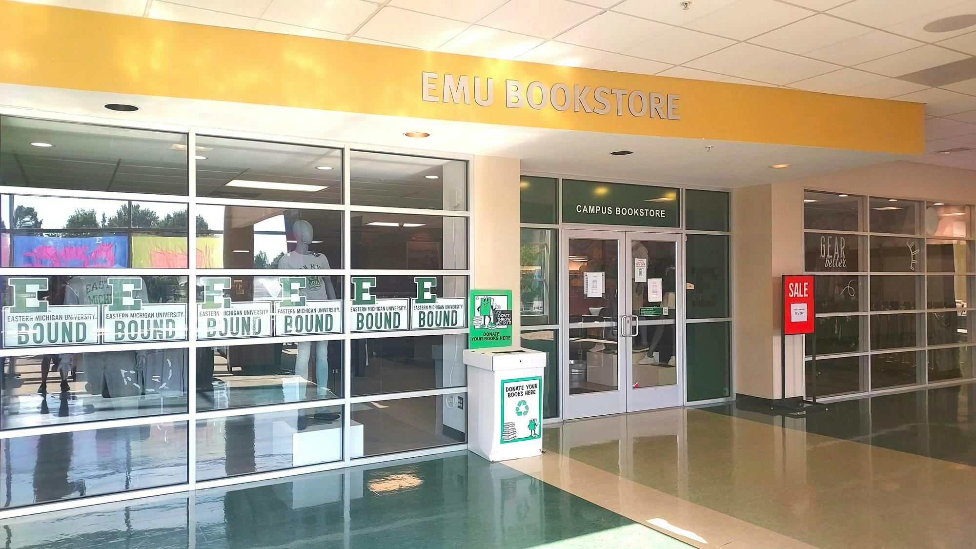 A wall with multiple windows leads to the campus bookstore. Multiple stickers on the window read "E Bound", and there is a donation box for books in front of the doors. 