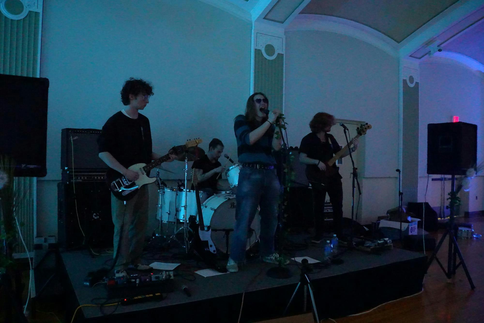 Four musicians performing onstage. Three musicians stand, while a fourth sits behind them playing the drums. The musicians standing on the far left and far right are playing guitar. The musician standing in the middle is wearing sunglasses and has their mouth open wide as they sing into the microphone they are holding.