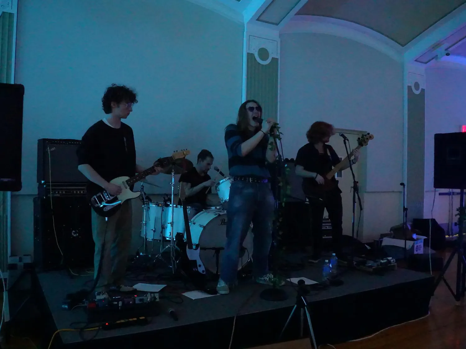 Four musicians performing onstage. Three musicians stand, while a fourth sits behind them playing the drums. The musicians standing on the far left and far right are playing guitar. The musician standing in the middle is wearing sunglasses and has their mouth open wide as they sing into the microphone they are holding.