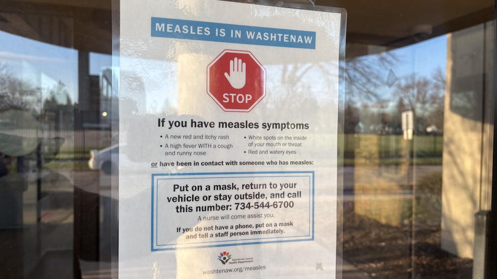 A sign on a glass door shows a stop sign symbol and says "Measles in Washtenaw." The sign instructs visitors to put on a mask, return to their vehicle or stay outside, and call 734-544-6700 if they are experiencing symptoms or have been around someone with measles.