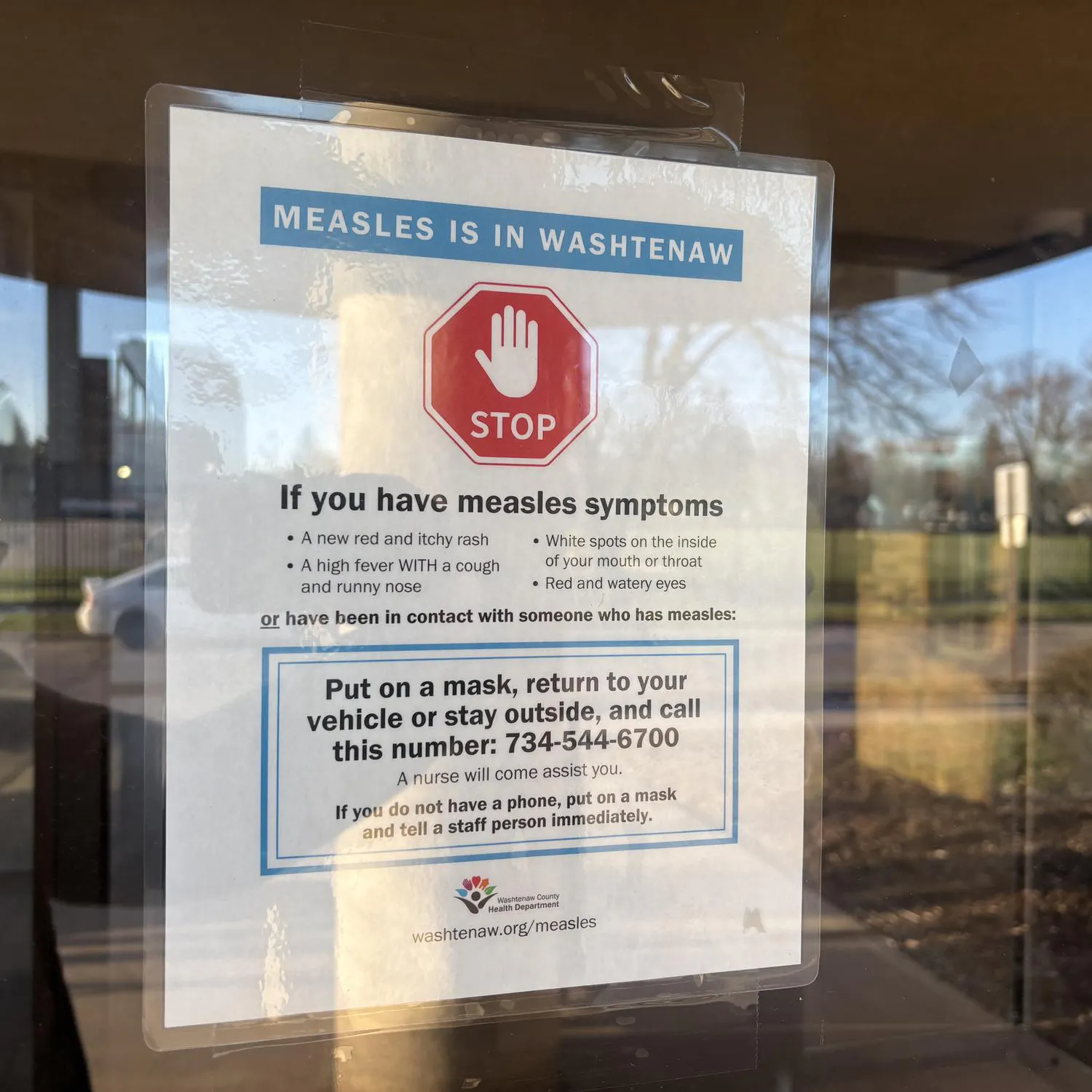 A sign on a glass door shows a stop sign symbol and says "Measles in Washtenaw." The sign instructs visitors to put on a mask, return to their vehicle or stay outside, and call 734-544-6700 if they are experiencing symptoms or have been around someone with measles.