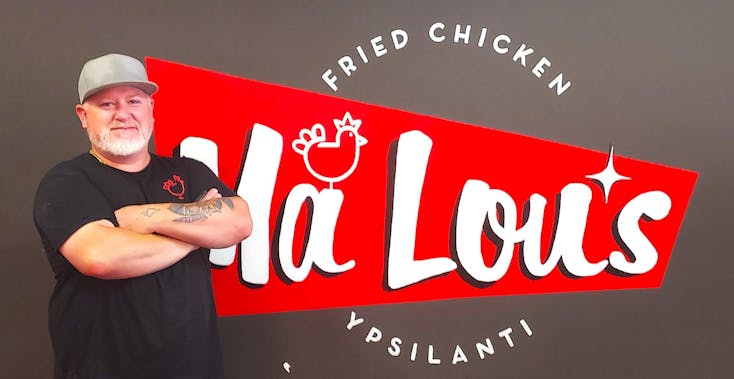 The restaurant owner has a grey hat, and he stands with his arms folded next to the Ma Lou's red-black-white colored logo wall. 