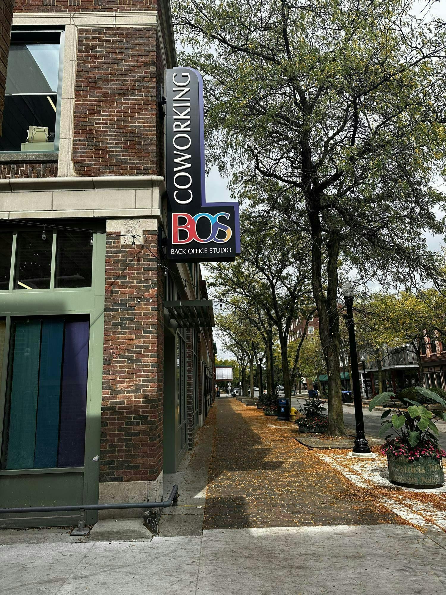 The edge of a brick building with a sign protruding from its right side. The sign is black and in the shape of the letter L. The top part of the sign says "Coworking" in white capital letters, and the word is written vertically. The bottom part of the sign says "BOS" in colorful capital letters, and beneath that, the words "Back Office Studio" are written in white capital letters. There is a row of trees with green leaves near the building, and the sky overhead is sunny.