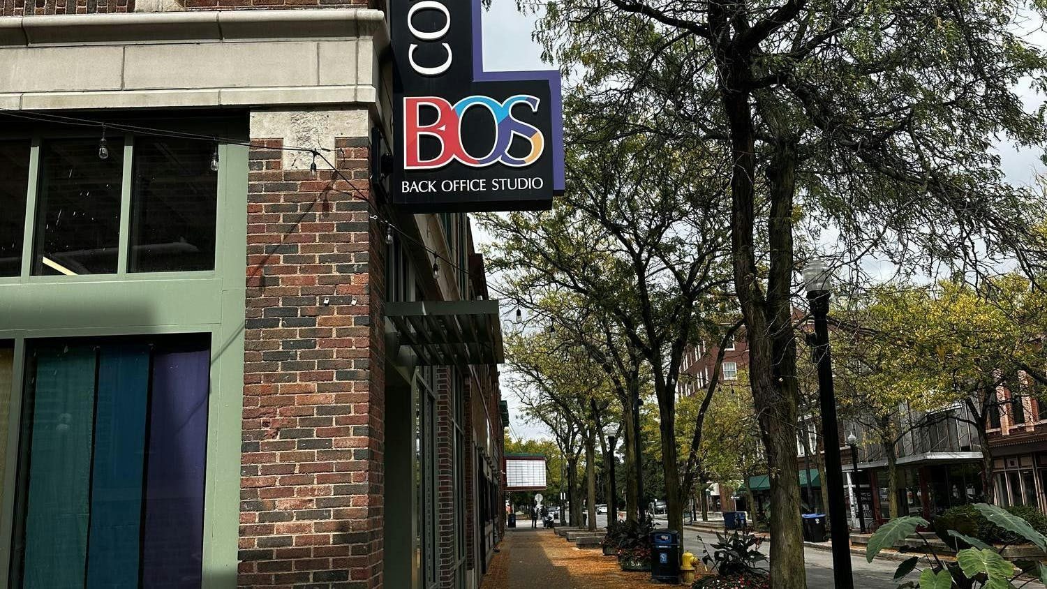 The edge of a brick building with a sign protruding from its right side. The sign is black and in the shape of the letter L. The top part of the sign says "Coworking" in white capital letters, and the word is written vertically. The bottom part of the sign says "BOS" in colorful capital letters, and beneath that, the words "Back Office Studio" are written in white capital letters. There is a row of trees with green leaves near the building, and the sky overhead is sunny.