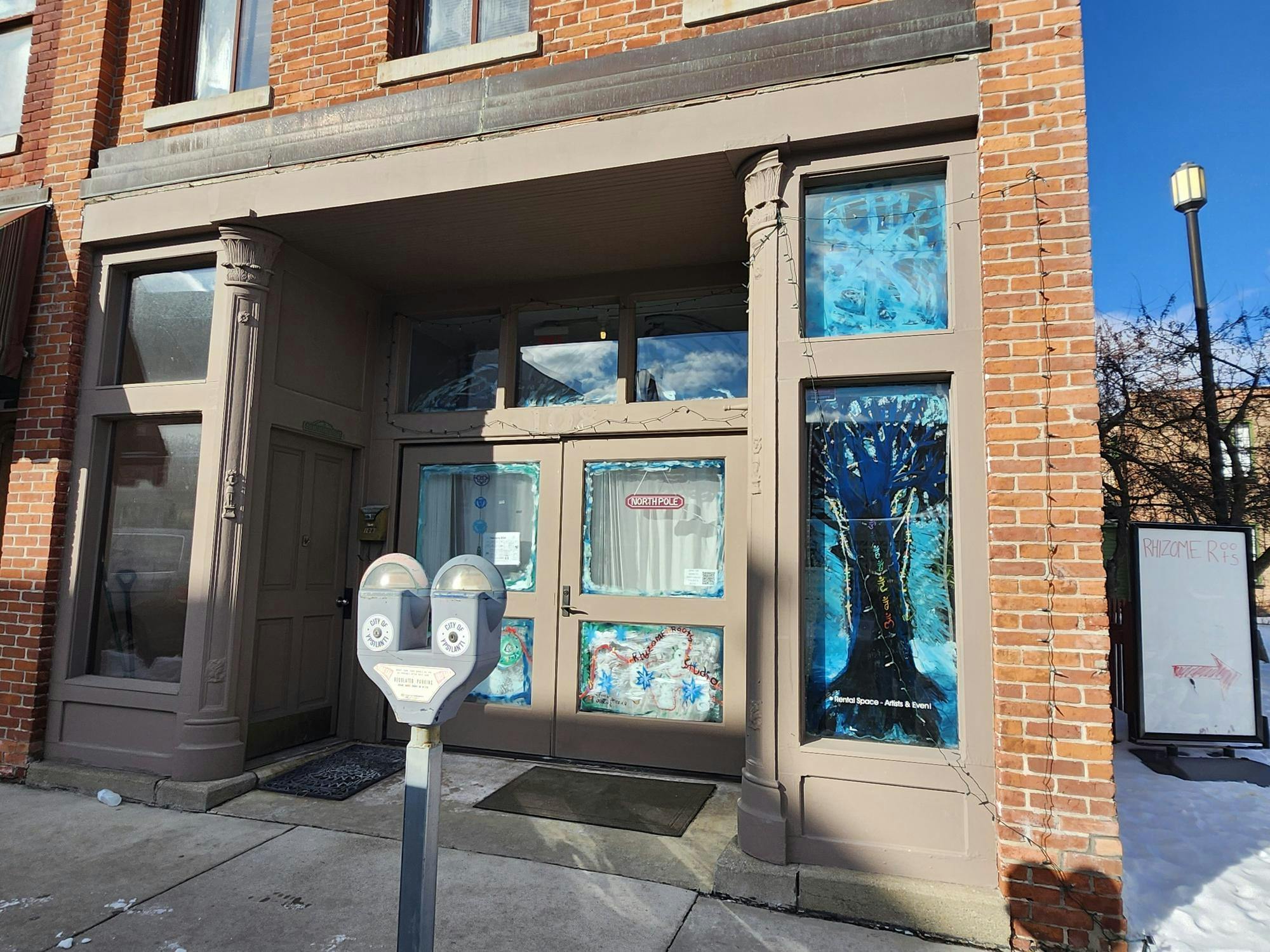 A brick building with a storefront with inset glass and wood paneled double doors. The windows on the doors and on either side of the doors are painted. There is an outline of a tree painted, as well as the text "Rhizome Roots Studio."