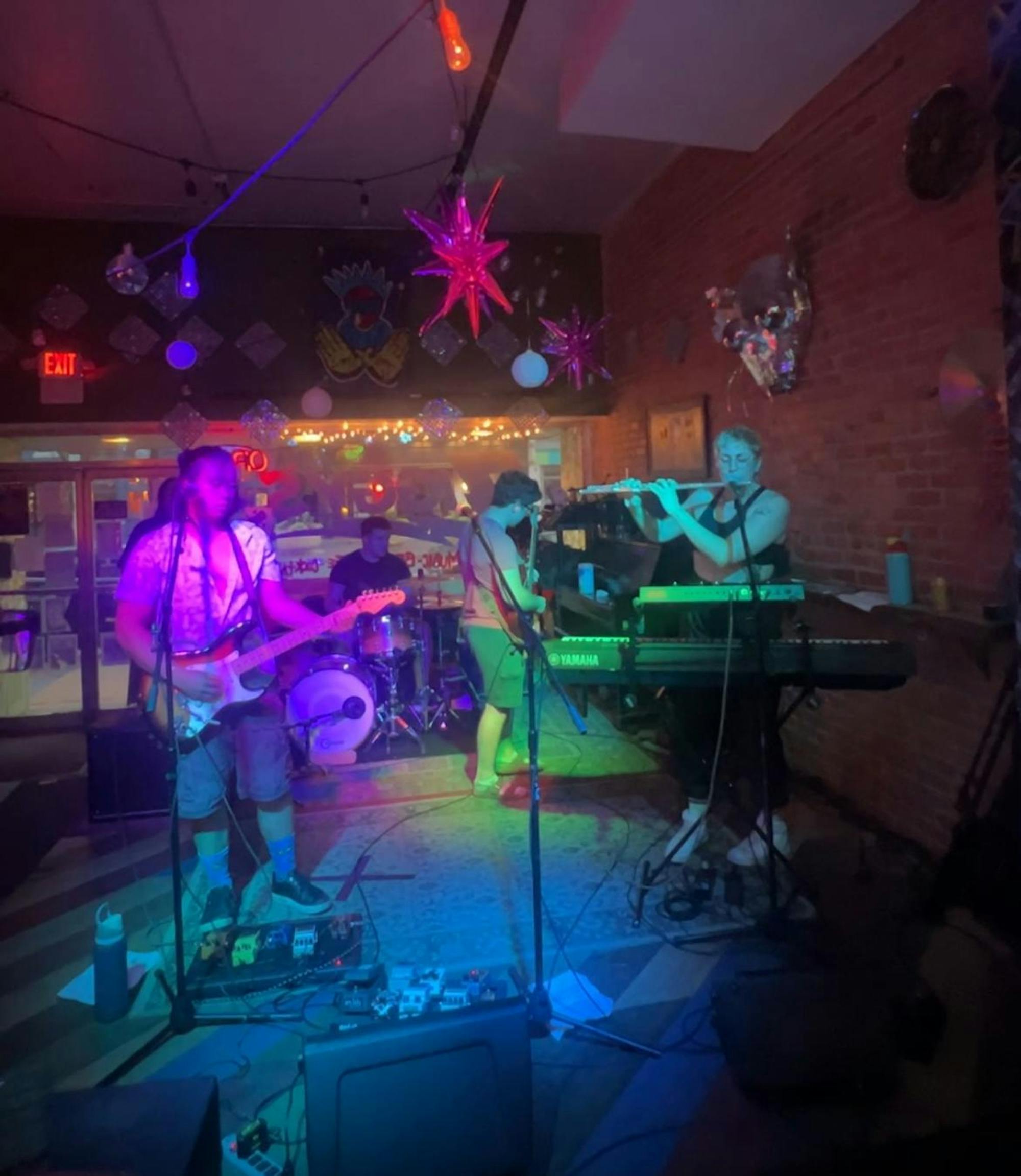 Members of a band on a colorfully lit stage playing flute, guitars, drums, and bass guitar. 