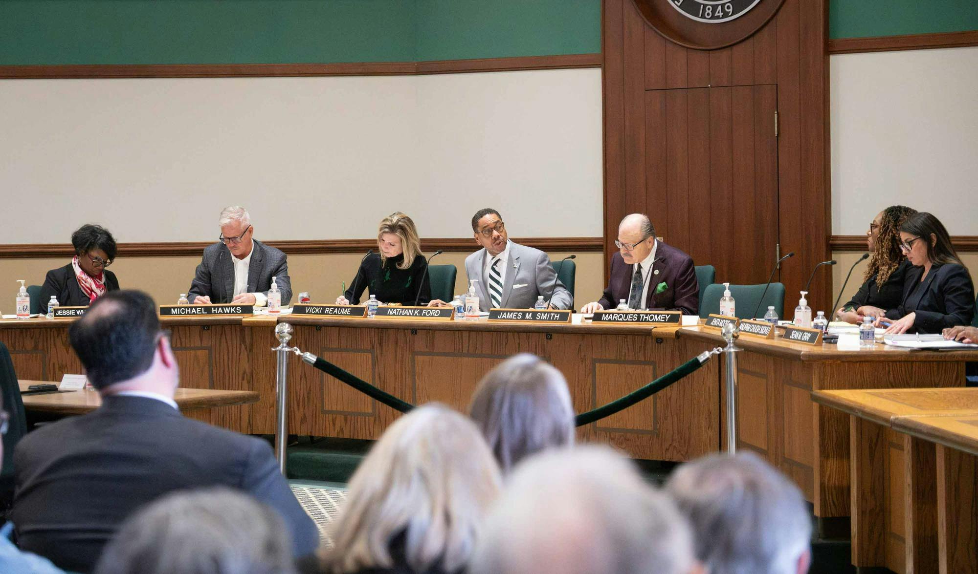 Regents and university officials sit at wooden desks. Attendees from the crowd are visible.