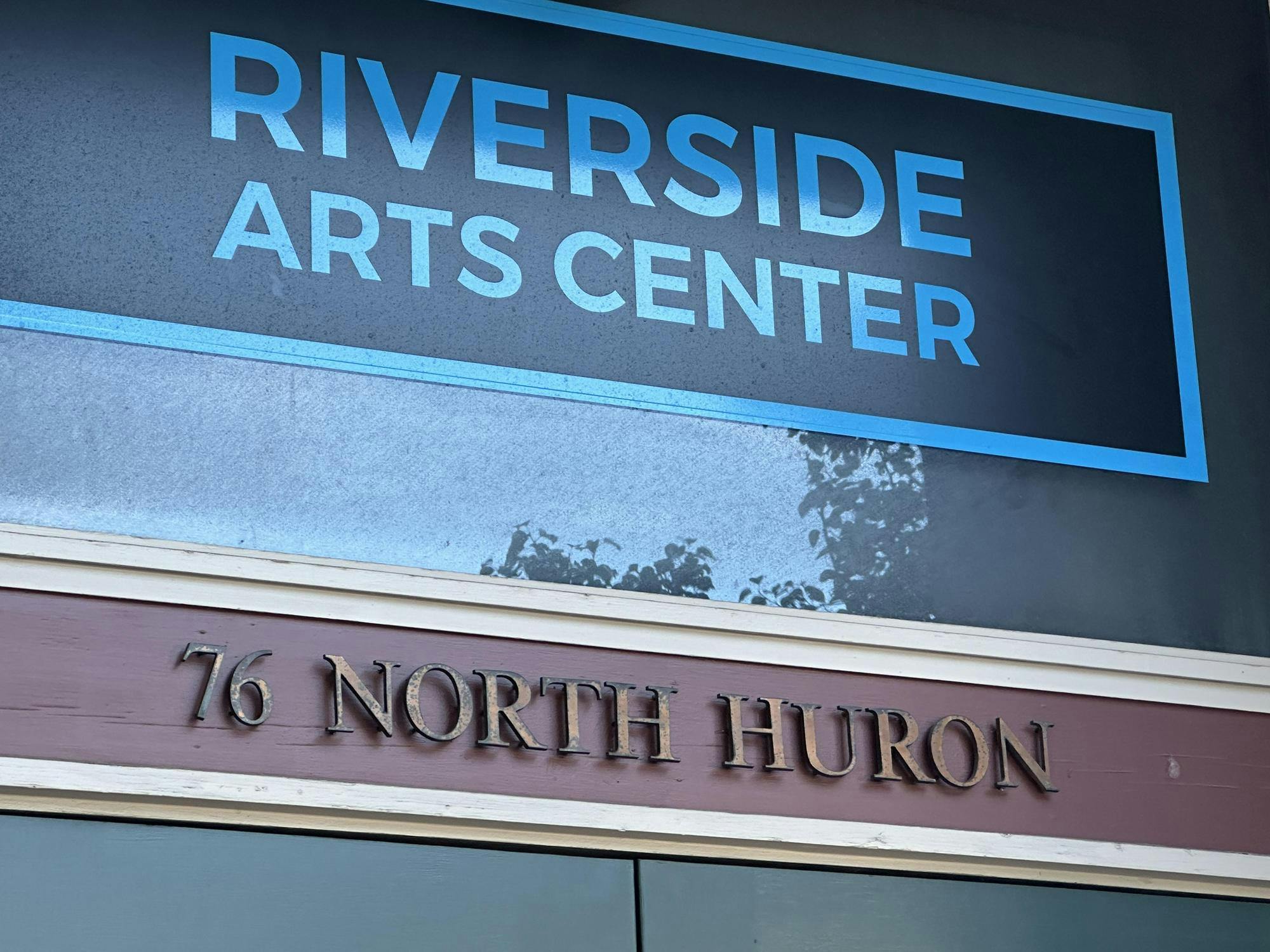 A close-up of the front of a building. At the top, big capital blue letters spell out "Riverside Arts Center" against a darker blue background. Underneath, raised rust-colored letters spell out "76 North Huron" against a maroon background. 