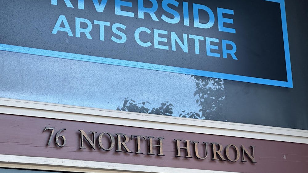 A close-up of the front of a building. At the top, big capital blue letters spell out "Riverside Arts Center" against a darker blue background. Underneath, raised rust-colored letters spell out "76 North Huron" against a maroon background.